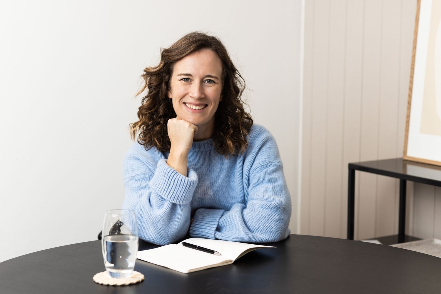 Professional in light blue sweater sitting at desk with notebook and water glass.