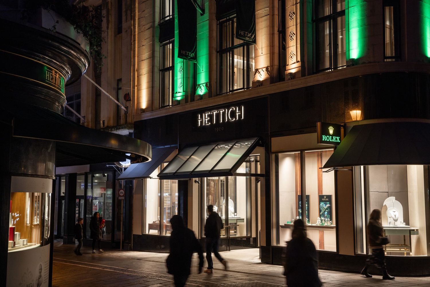 Hettich storefront at night with illuminated windows and pedestrians walking past.