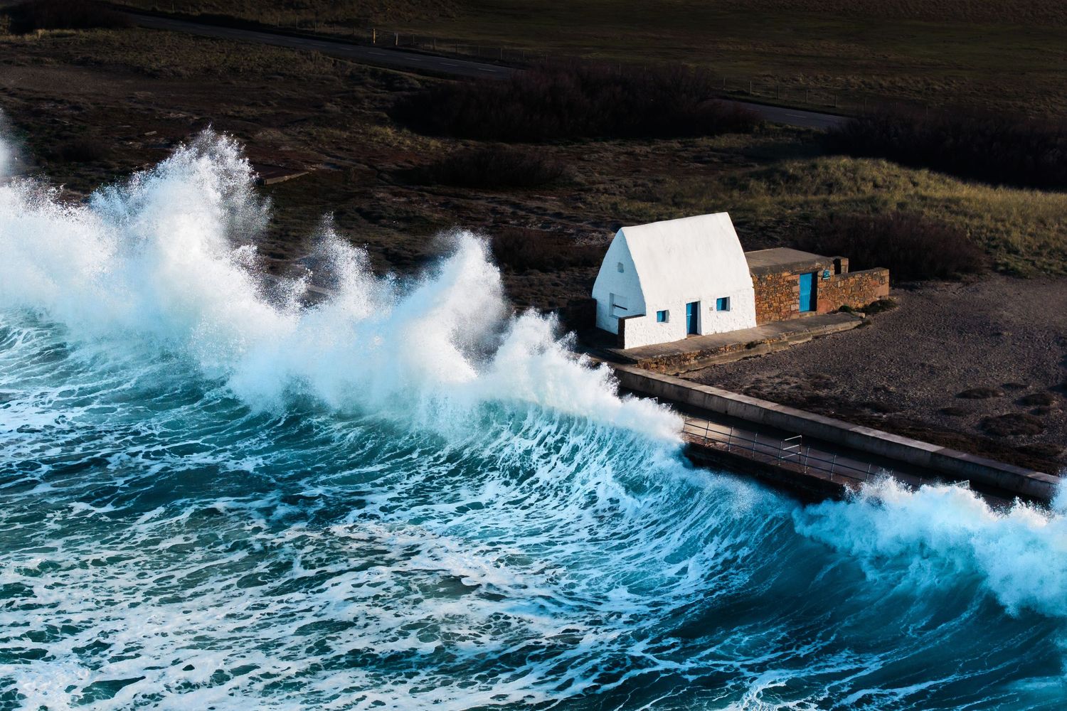 Massive blue waves crash against a small white house perched on a rocky coastal cliff.