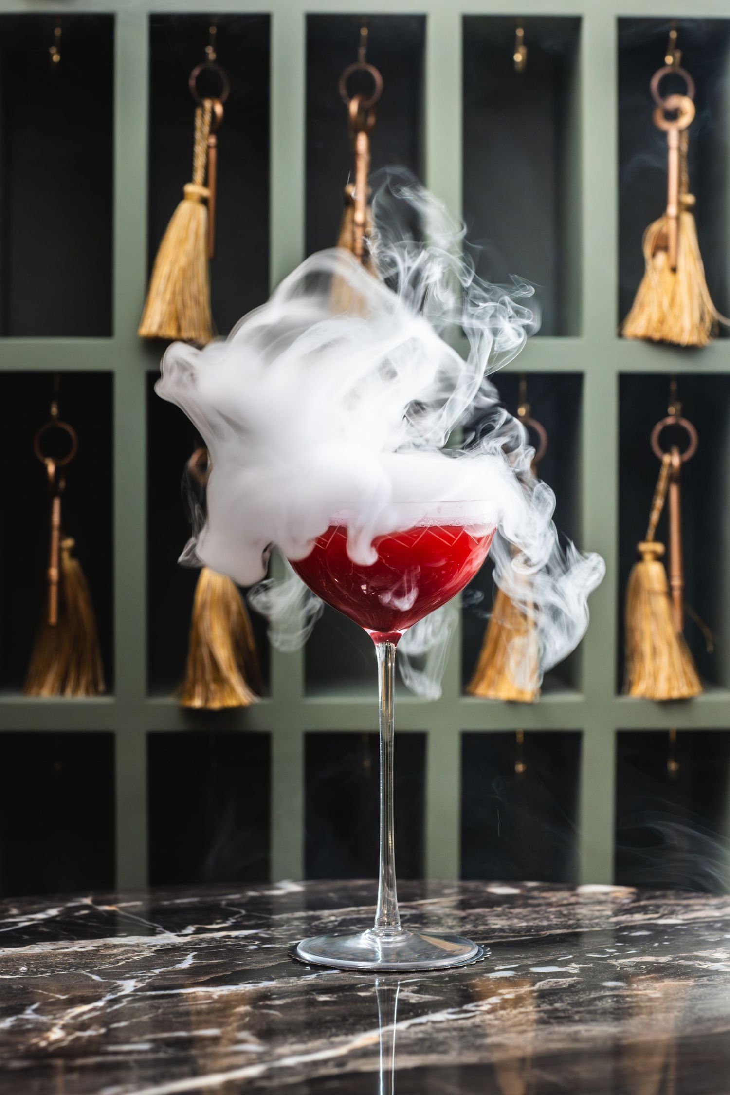 A dramatic red cocktail with white smoke effect against a backdrop of hanging gold tassels.