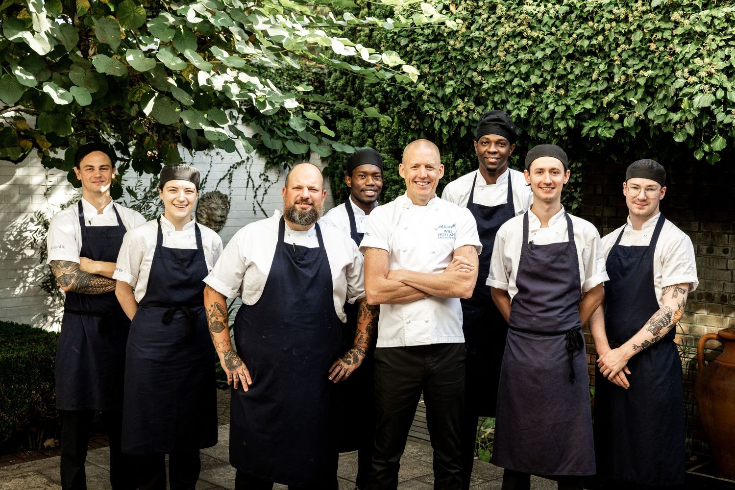 Professional kitchen staff posing together in front of a green hedge backdrop.
