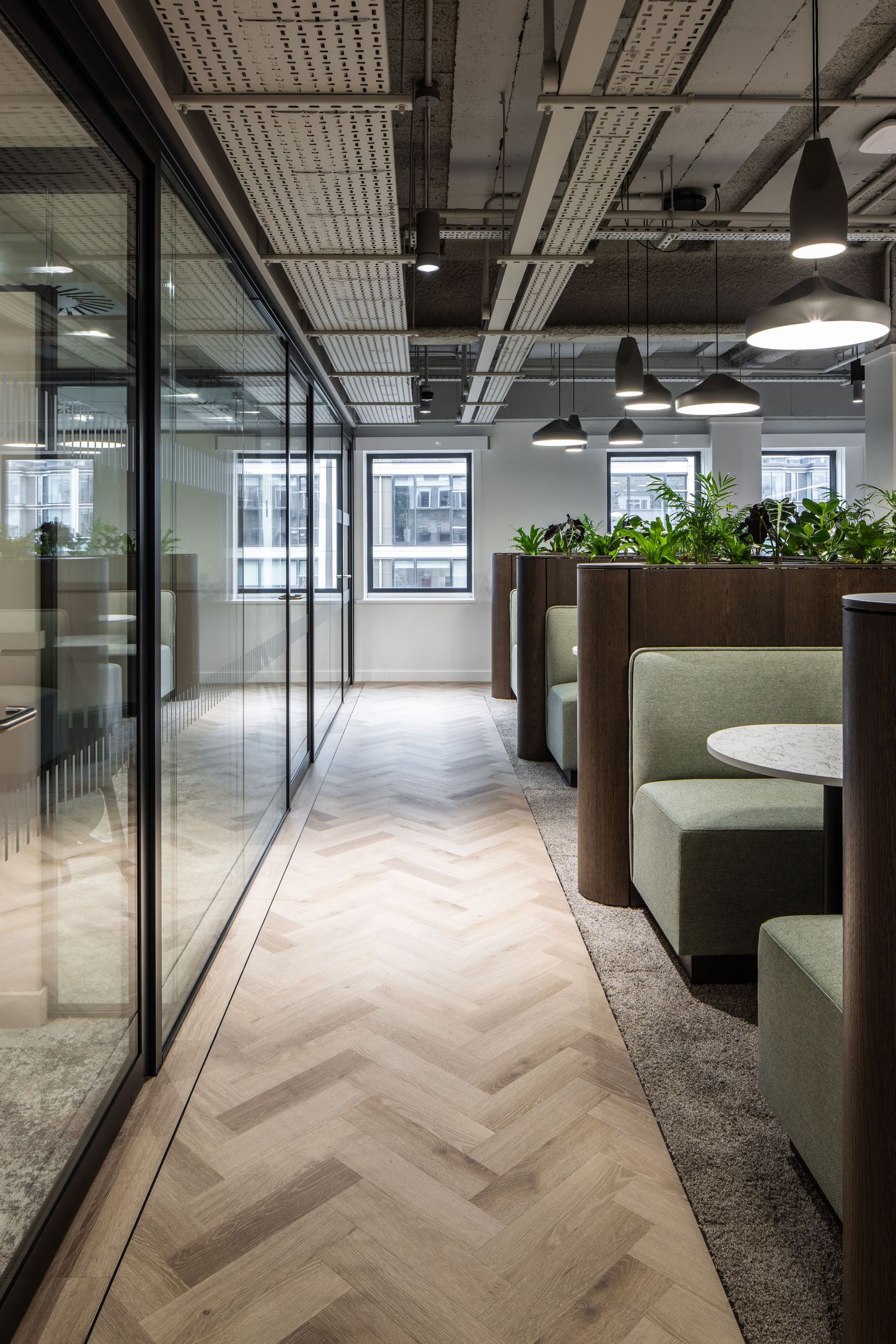 Long office corridor with glass walls, indoor plants, and chevron patterned flooring leading to bright windows.