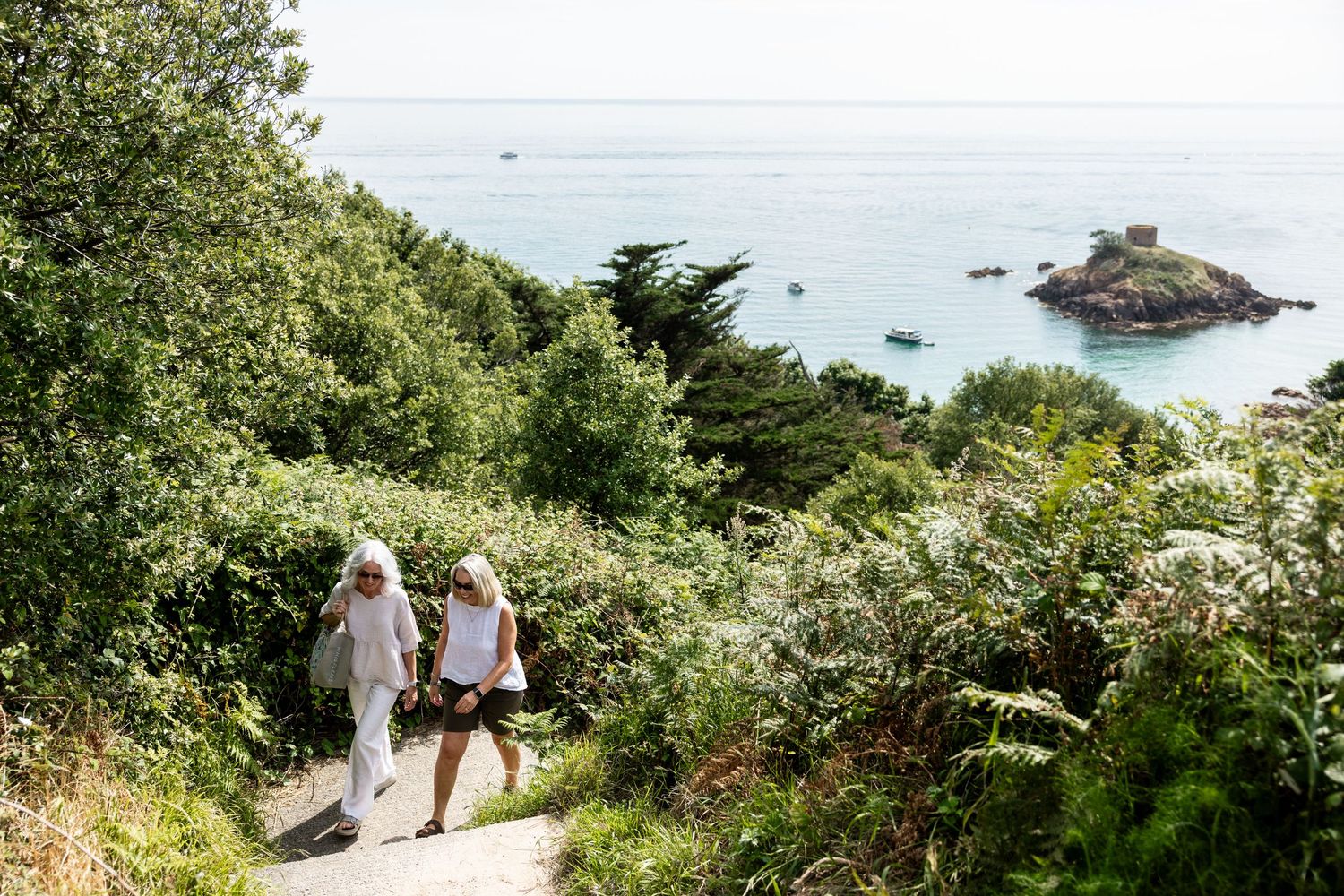 Two people walking down scenic coastal path with ocean views and historic fortress in distance.