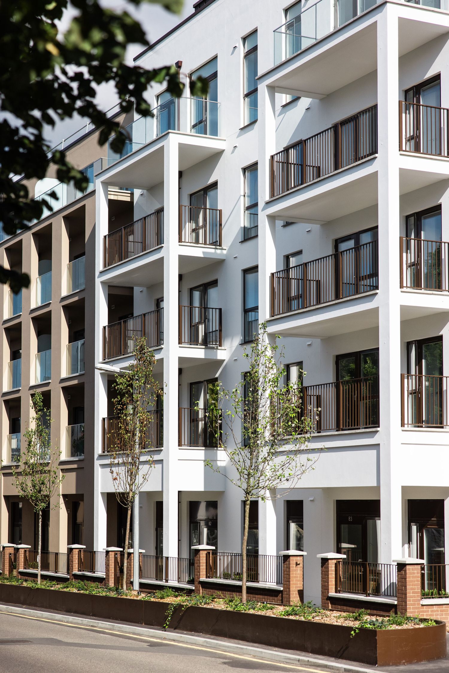 White modern apartment building with repeating balconies and glass railings along a landscaped street.