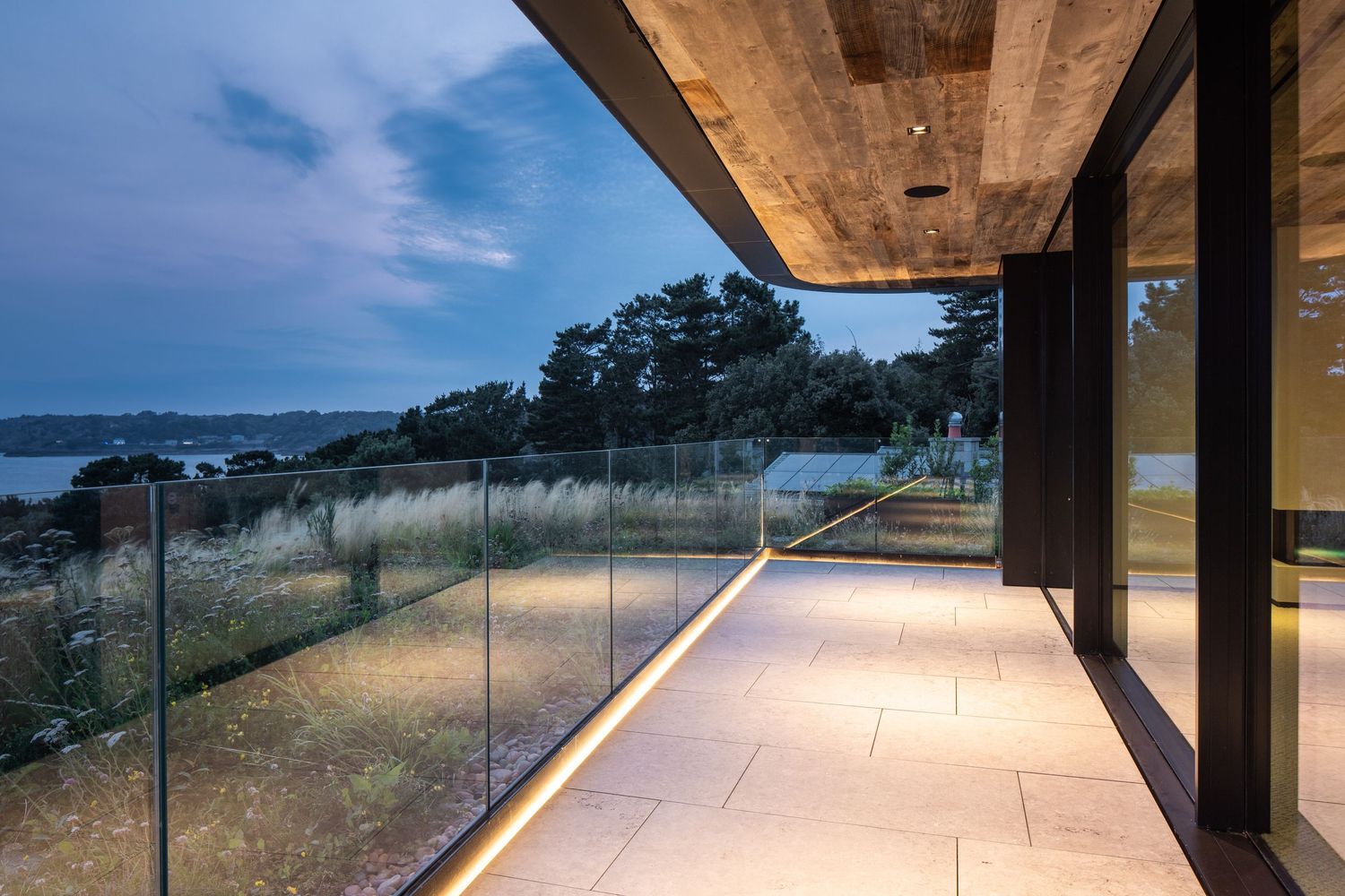 Modern balcony with glass railings overlooking coastal landscape at dusk with LED floor lighting.