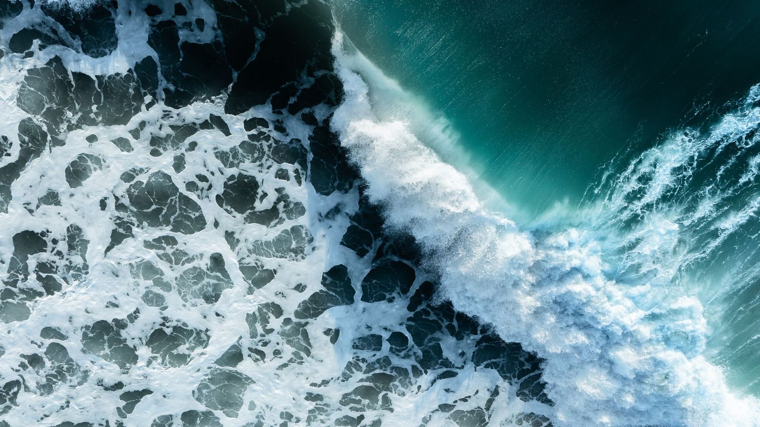 Dramatic aerial view of powerful ocean waves crashing against a rocky coastline with white foam spray.