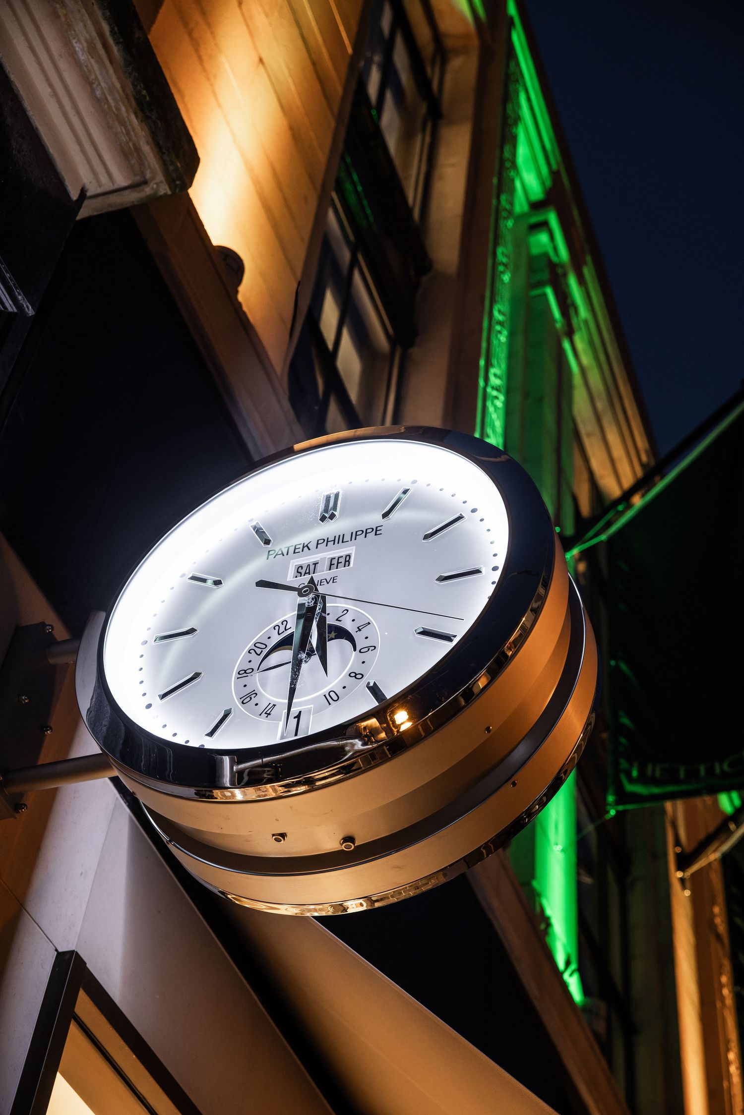 Large mounted clock on building exterior with green lighting in background.