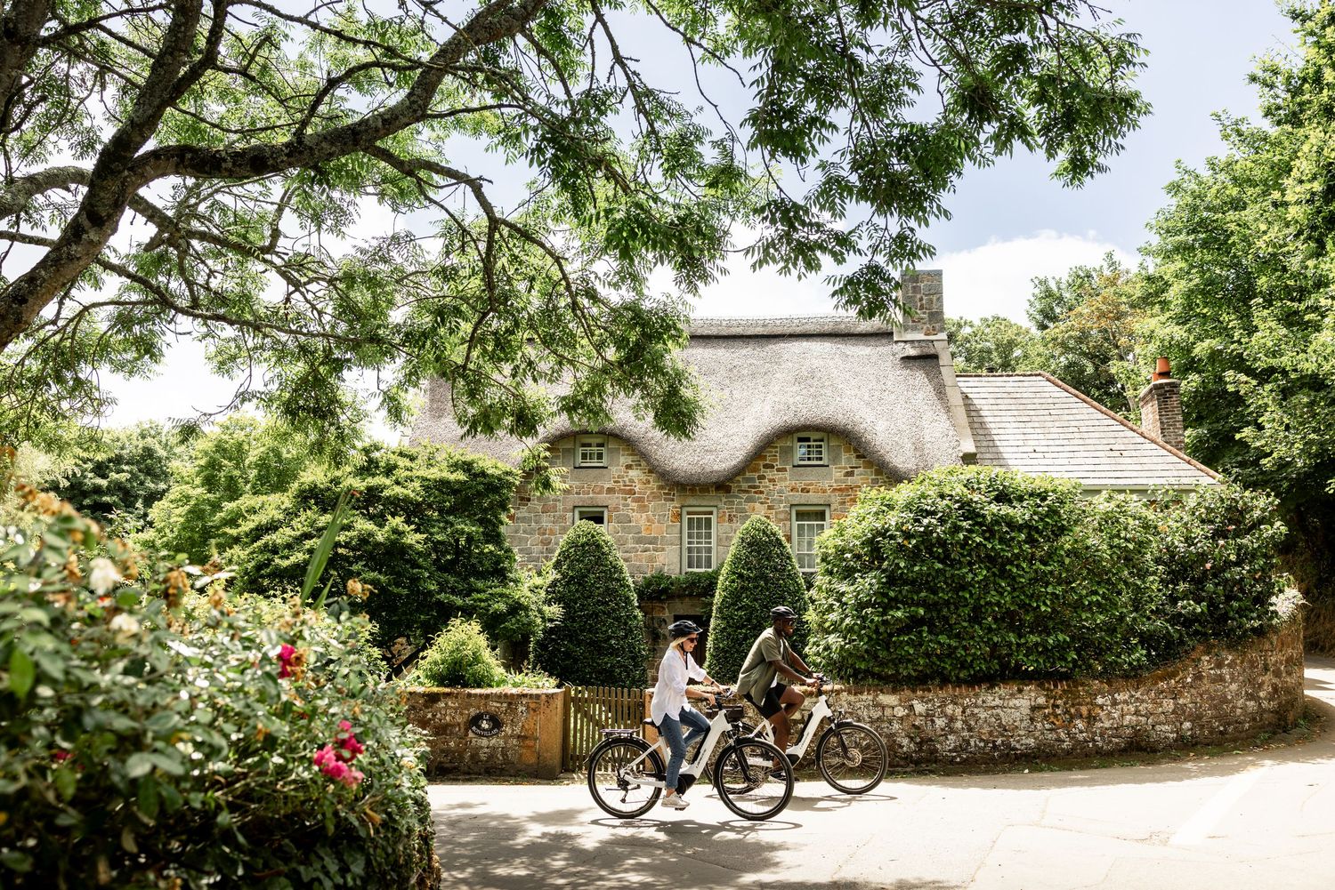 A cyclist rides past a charming thatched-roof cottage with stone walls and climbing vines in a lush garden setting.
