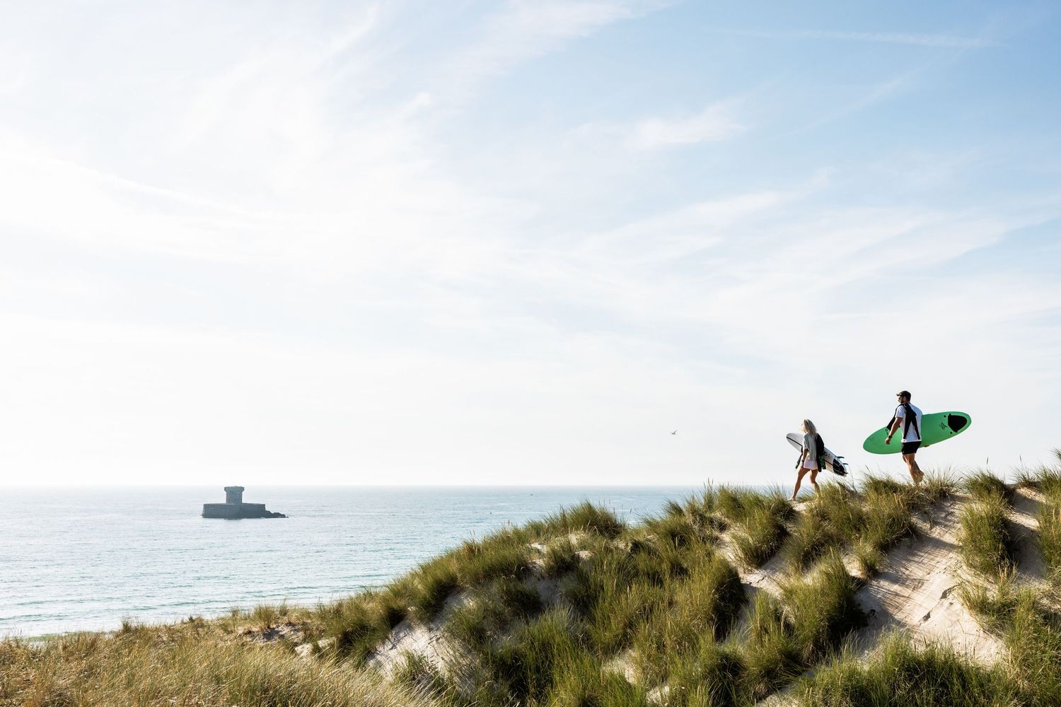 Surfers walking along sandy dunes with surfboards against a bright blue sky.