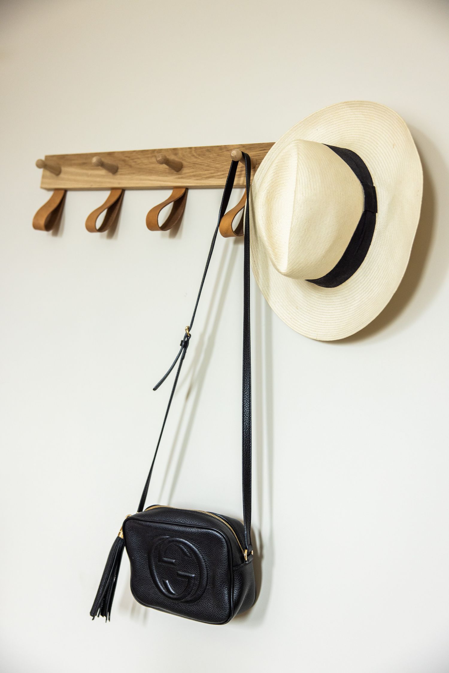Minimalist white wall decor with wooden coat hooks displaying a black handbag and cream-colored straw hat.