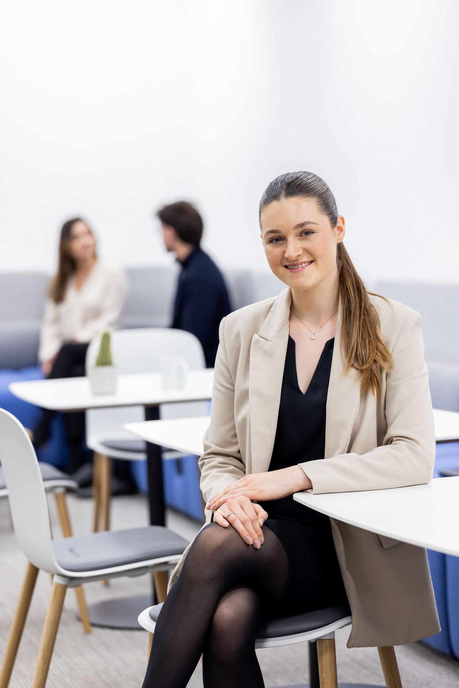 Professional in beige blazer sits casually on chair in modern office setting.