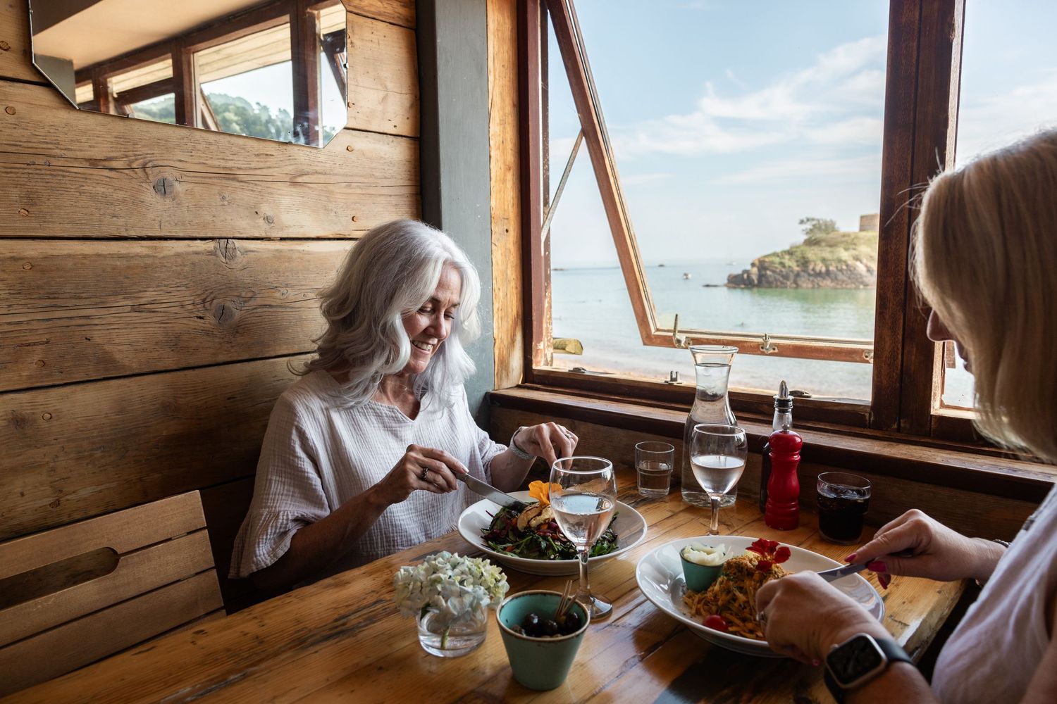 Two people enjoying a meal at a rustic waterfront restaurant with scenic ocean views through wooden windows.