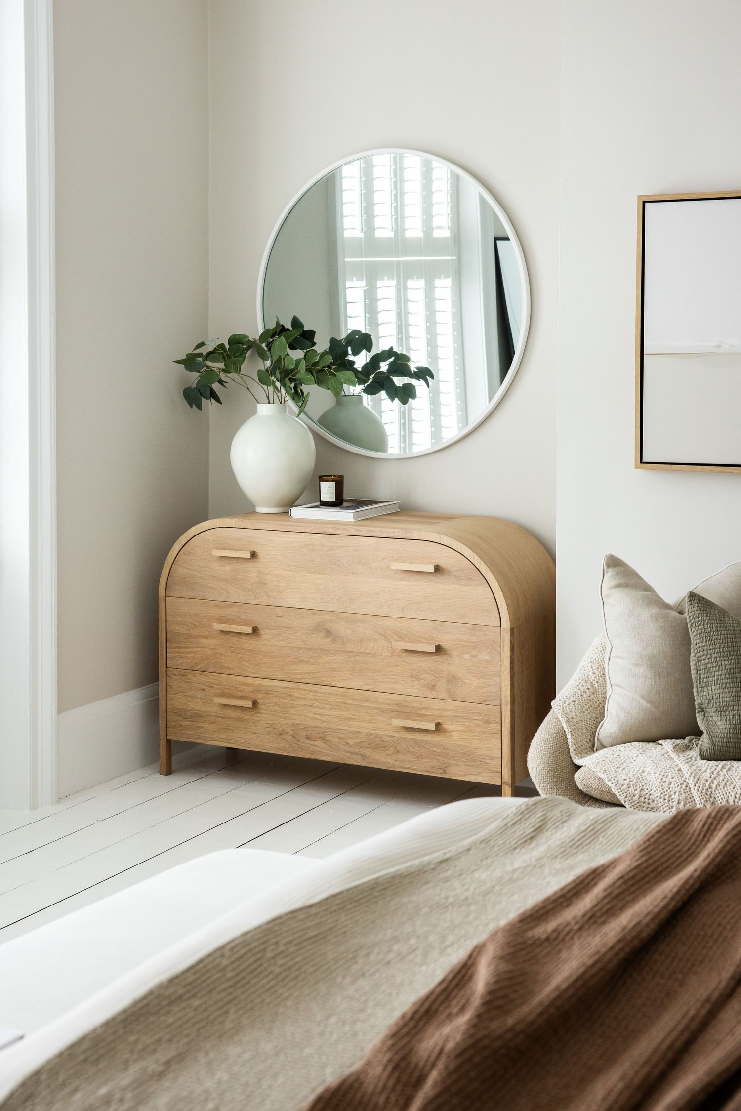 Wooden dresser with curved edges styled with round mirror and greenery in a serene bedroom corner.