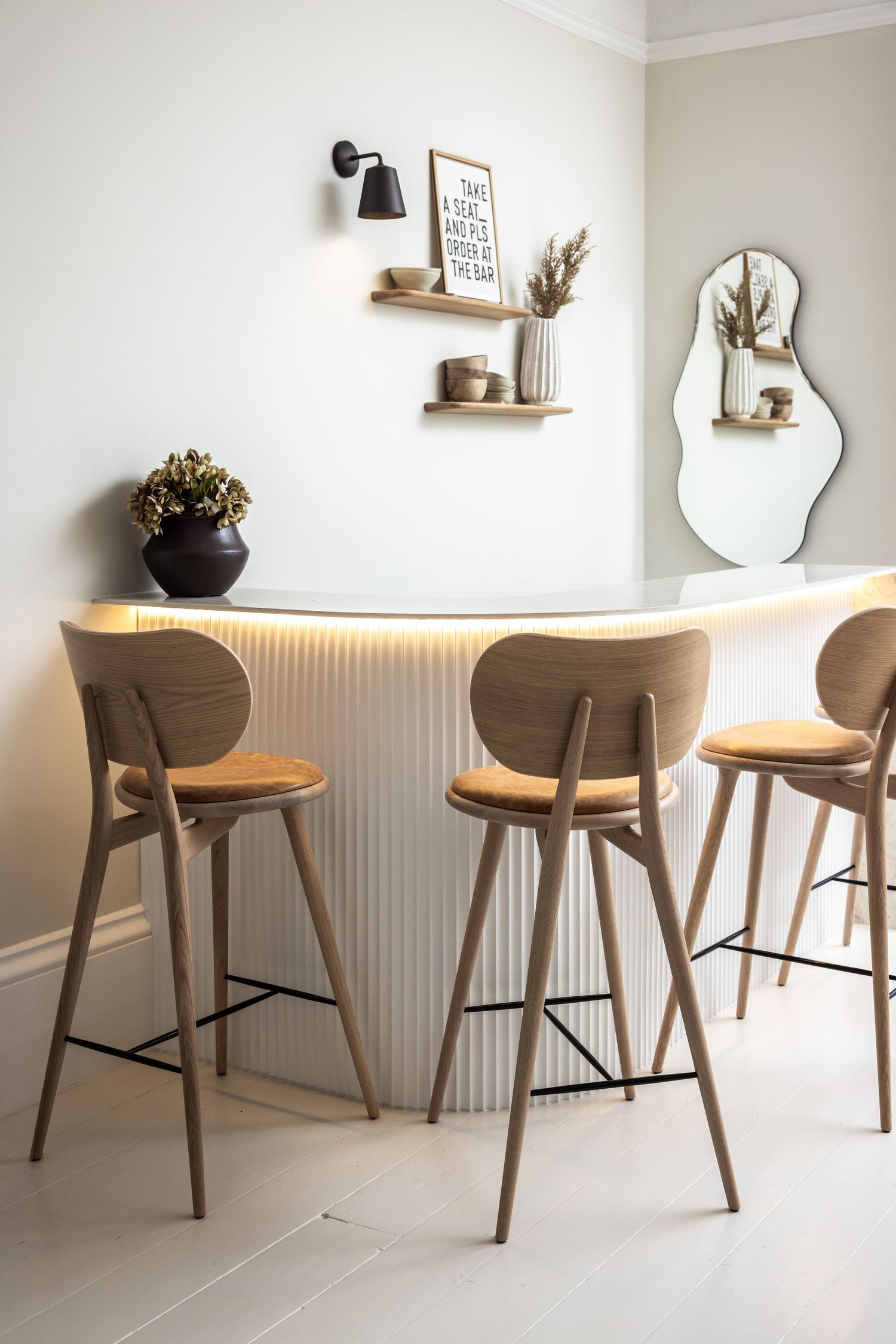 Modern bar seating area with wooden stools, floating shelves, and a decorative mirror in a minimalist white interior.