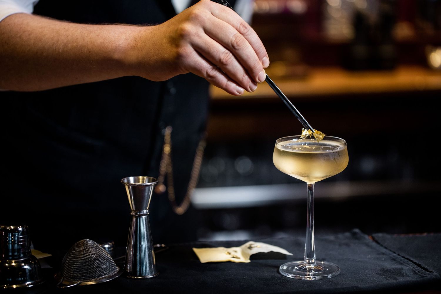 Bartender garnishing a golden cocktail in coupe glass at dark bar counter.