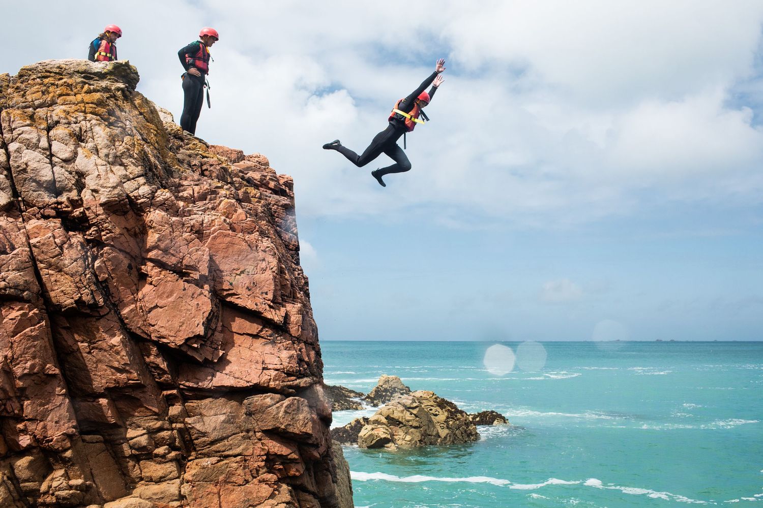 Person leaping off rocky cliff into turquoise ocean waters while others watch from above.