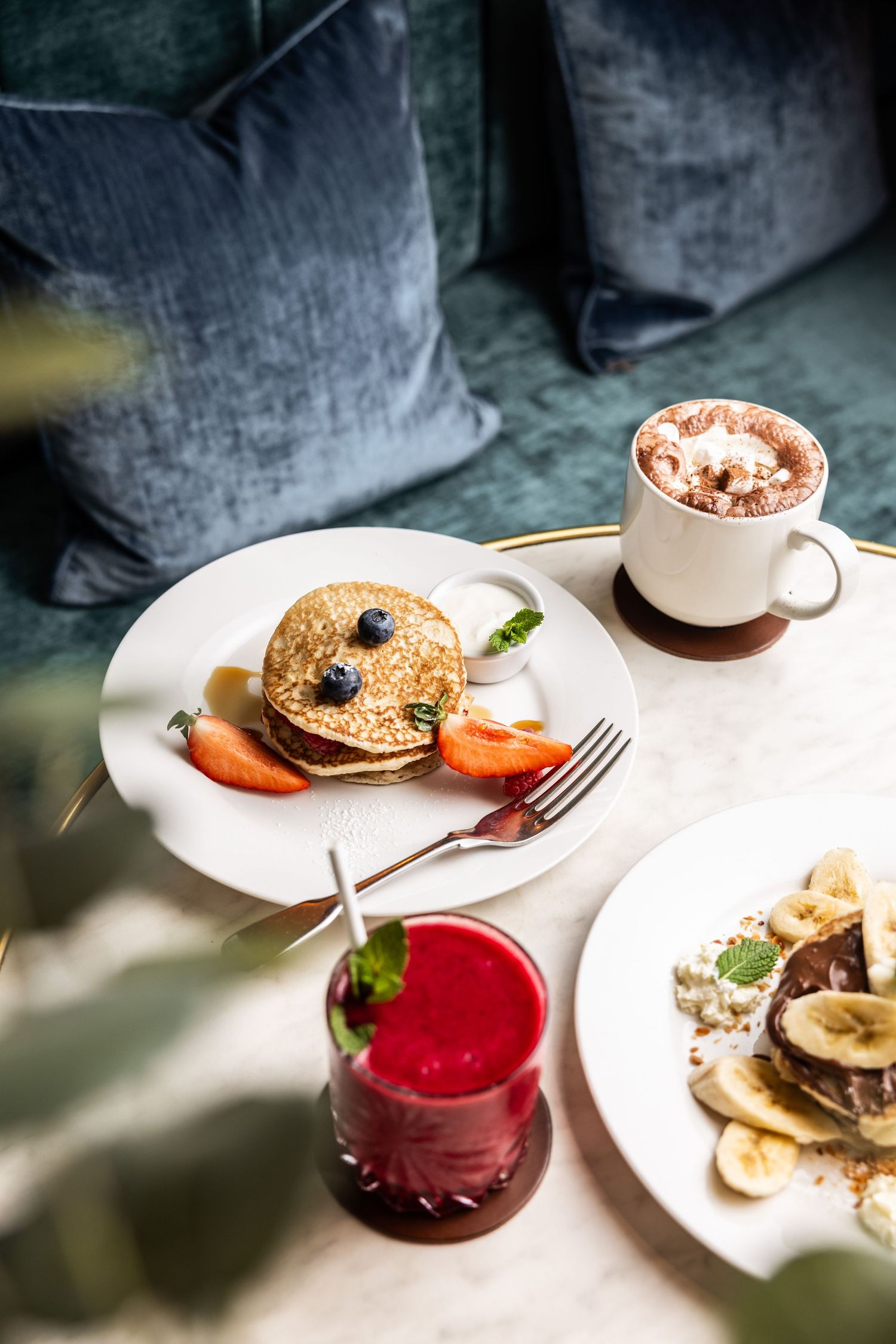 Breakfast spread with pancakes, red juice, and fresh fruits on white plates against a dark background.