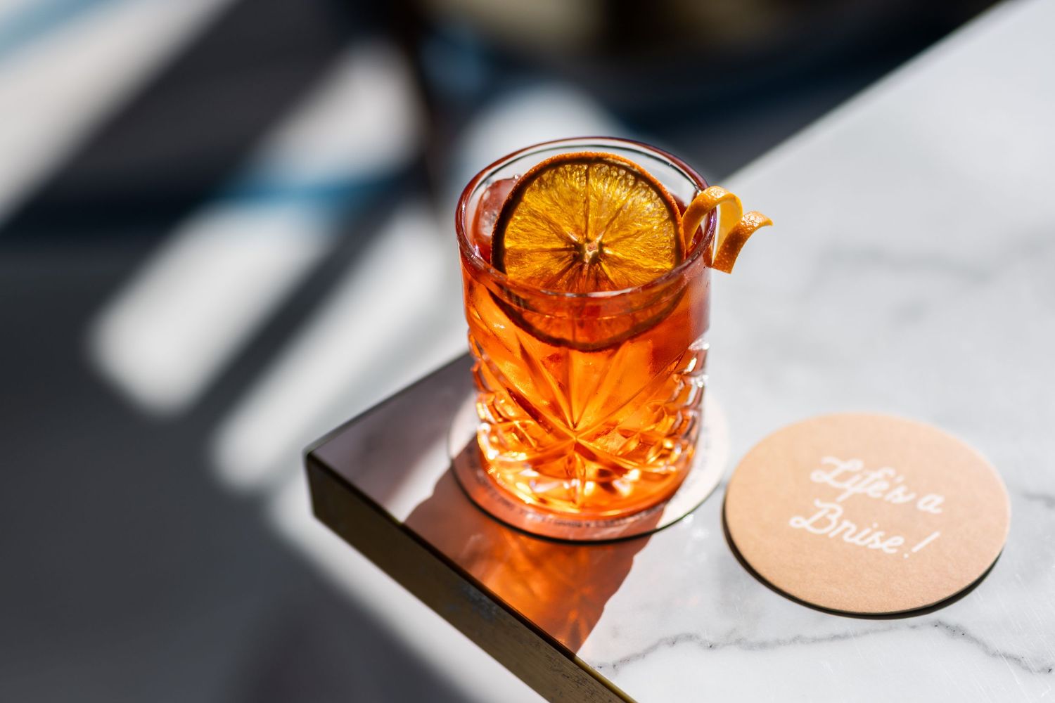 Orange cocktail with citrus garnish in a crystal glass catching sunlight on a marble surface.