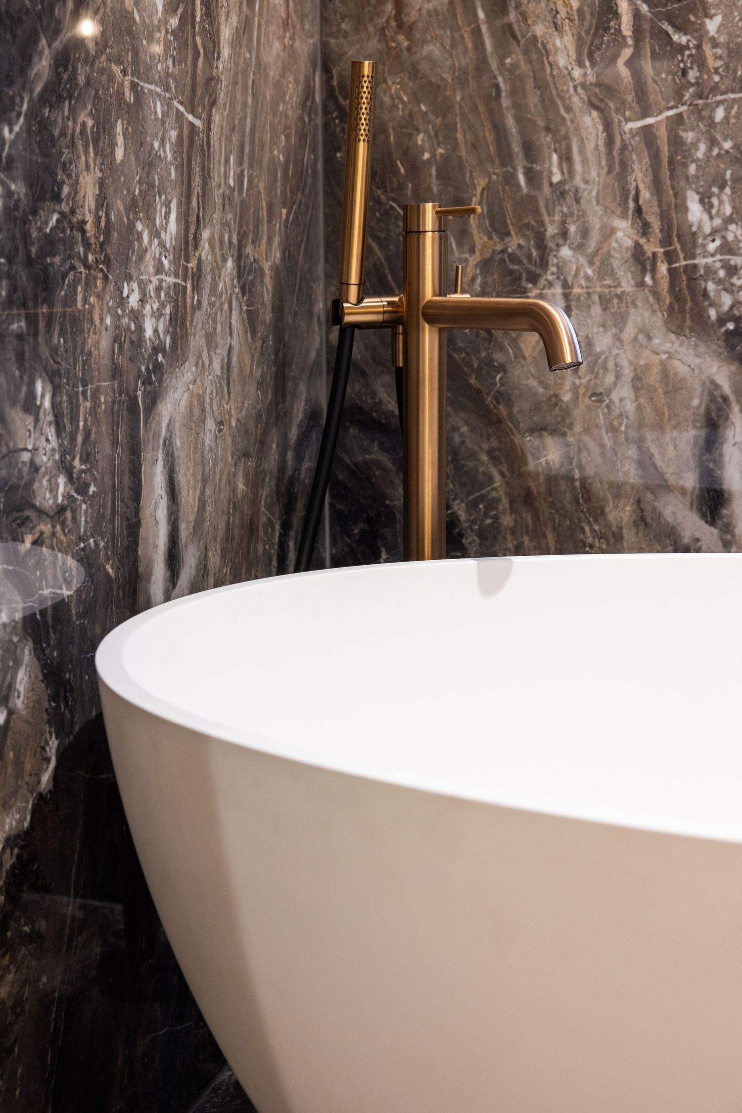 Modern freestanding white bathtub with gold faucet fixture against dark marble wall in luxury bathroom.