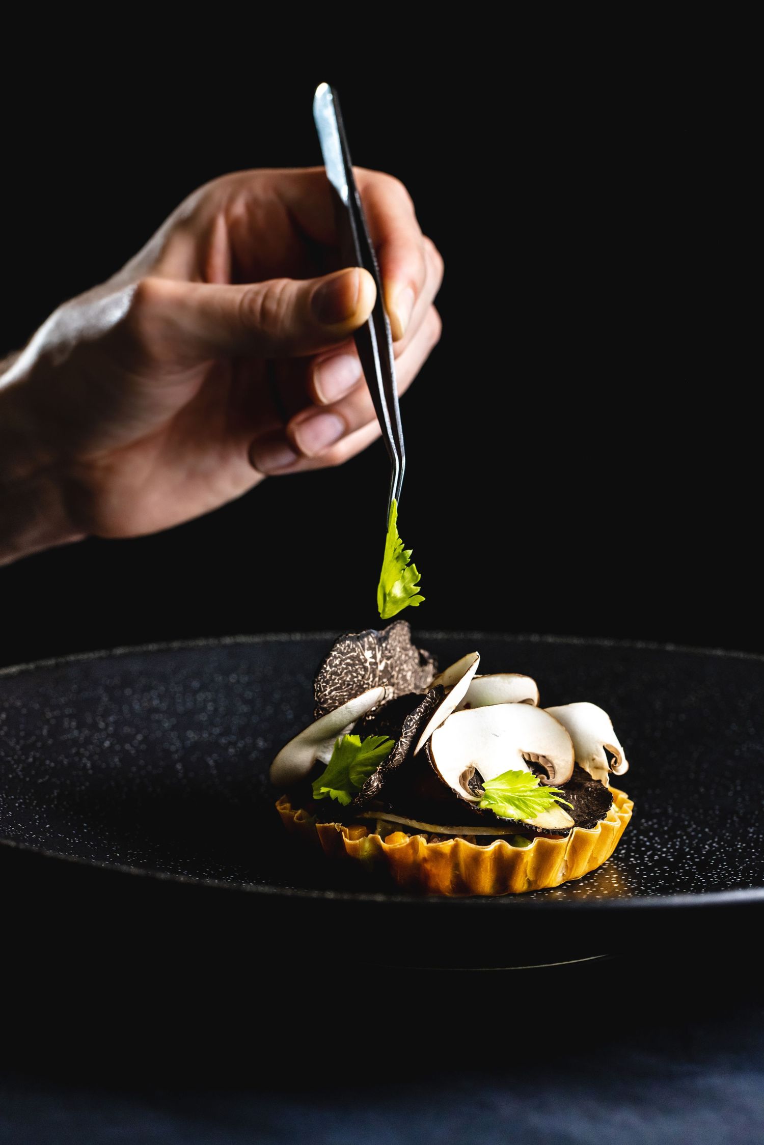 Hand placing garnish on an elegant black tart with mushrooms and microgreens.