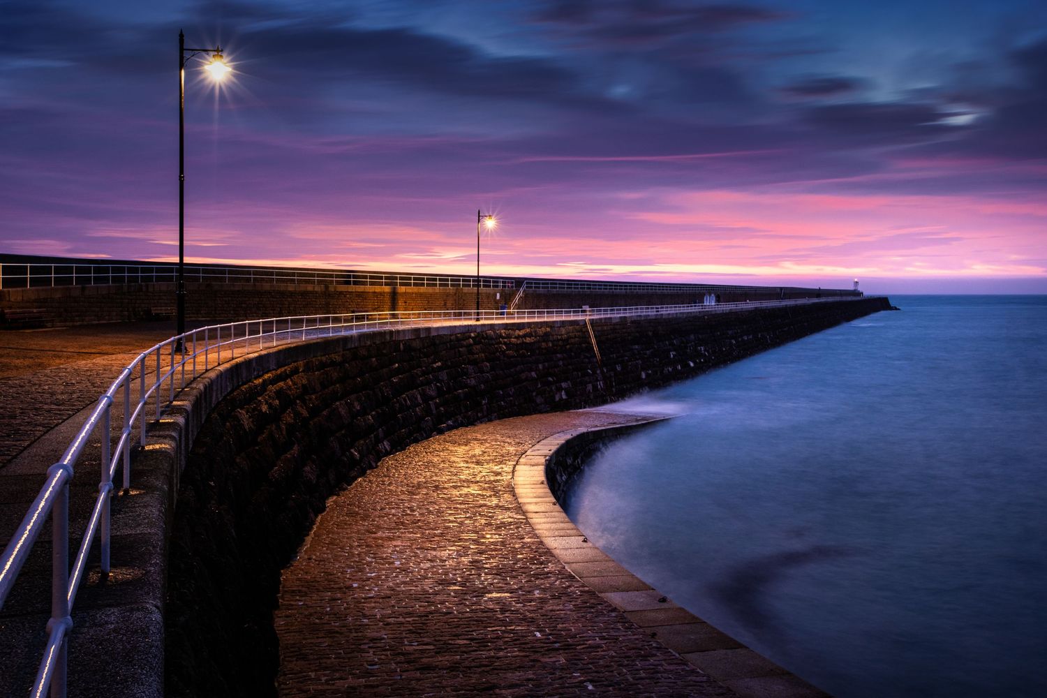 Curved seaside walkway with lampposts illuminated against a deep purple twilight sky.