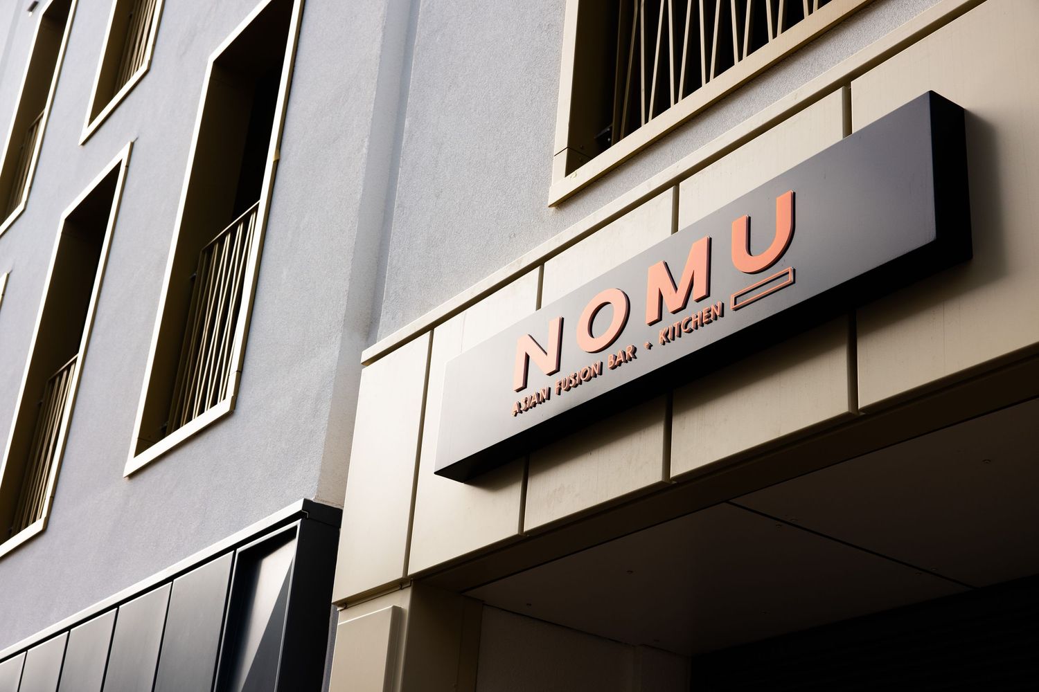 Close-up of illuminated Nomu signage mounted on beige building exterior.