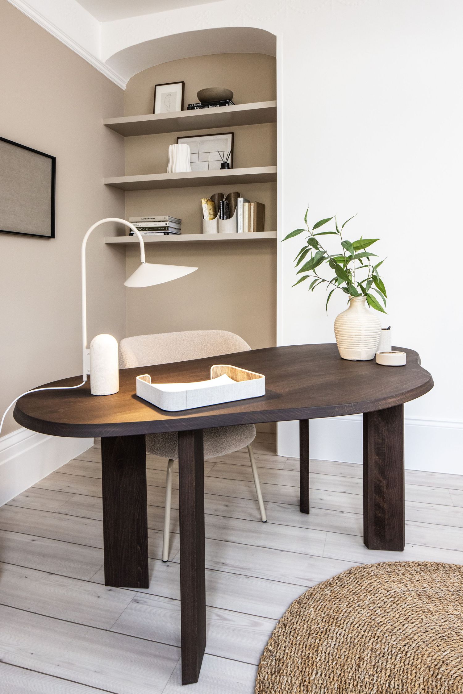 Modern dark wood desk with curved edges sits on light wood flooring against a beige wall with floating shelves.