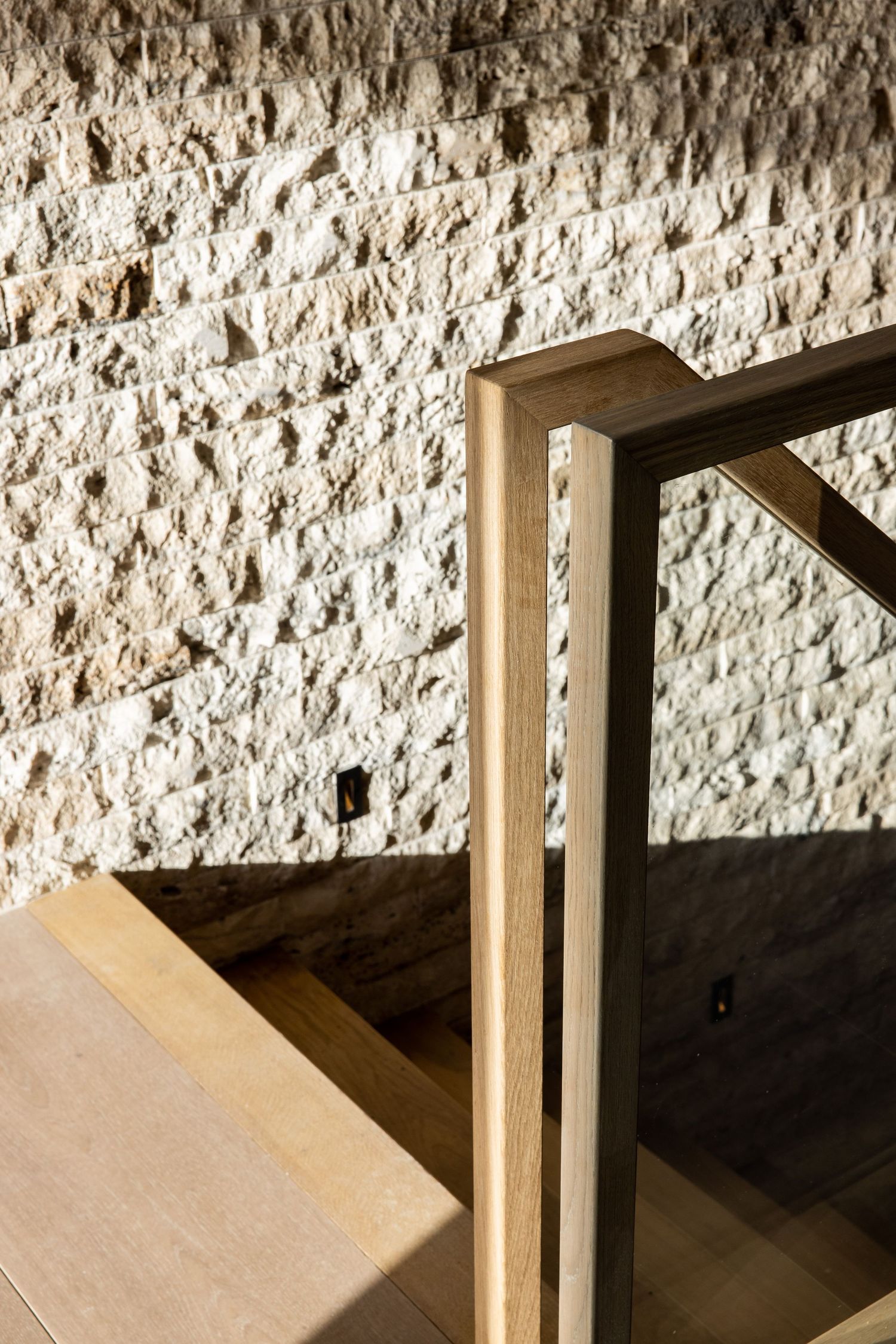 Modern wooden staircase detail against textured white stone wall with dramatic lighting and shadows.