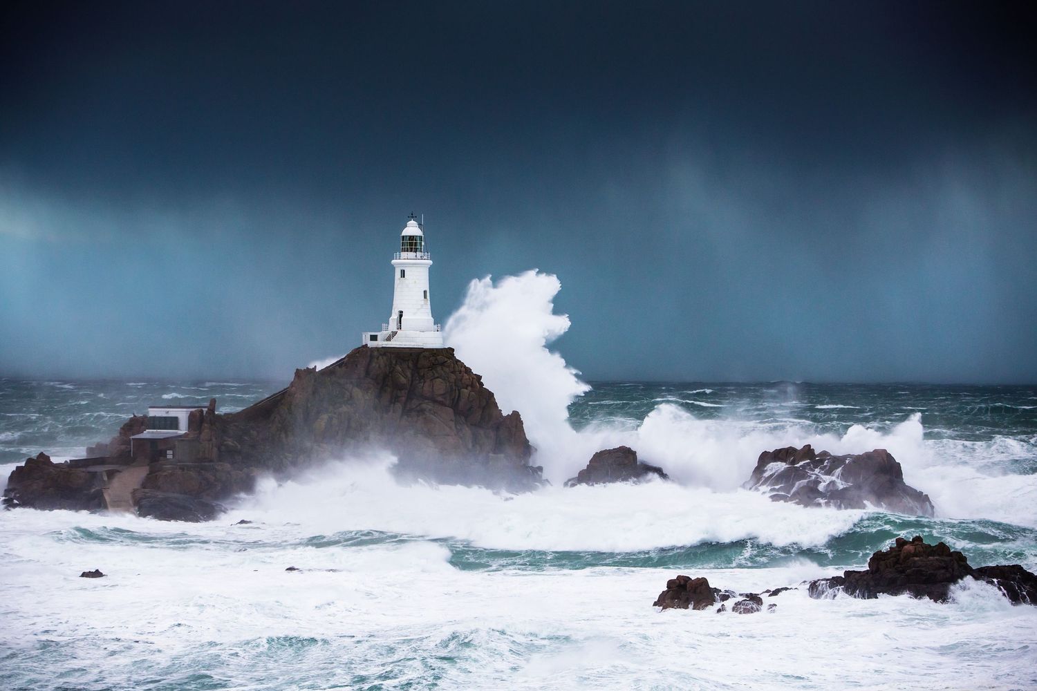 Powerful ocean waves crashing against rocky outcrops near a lighthouse in stormy weather.