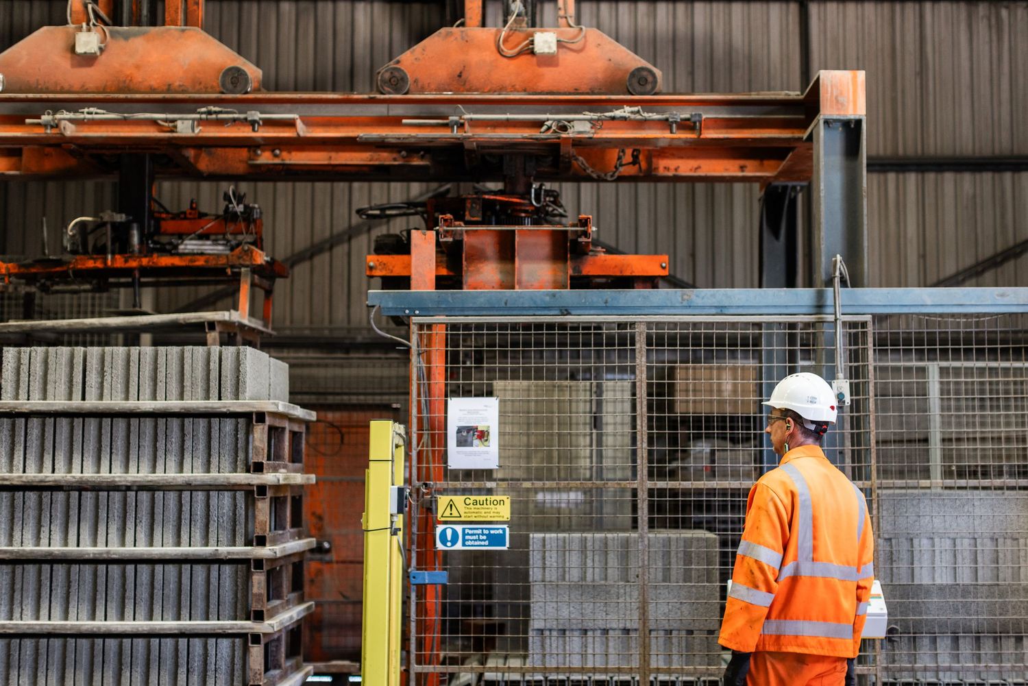 Workers in orange safety vests operate heavy machinery in an industrial warehouse setting.