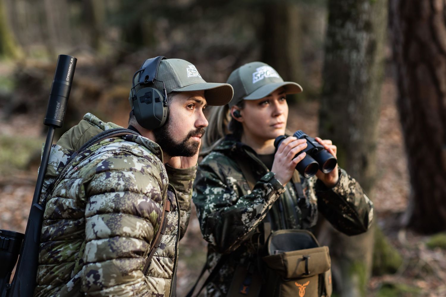 Two hunters in camouflage gear and caps scout the woods, one holding binoculars and the other carrying a rifle.
