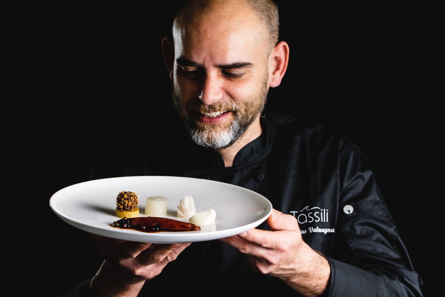 Plating a gourmet dish with tweezers on a white plate against dark background.