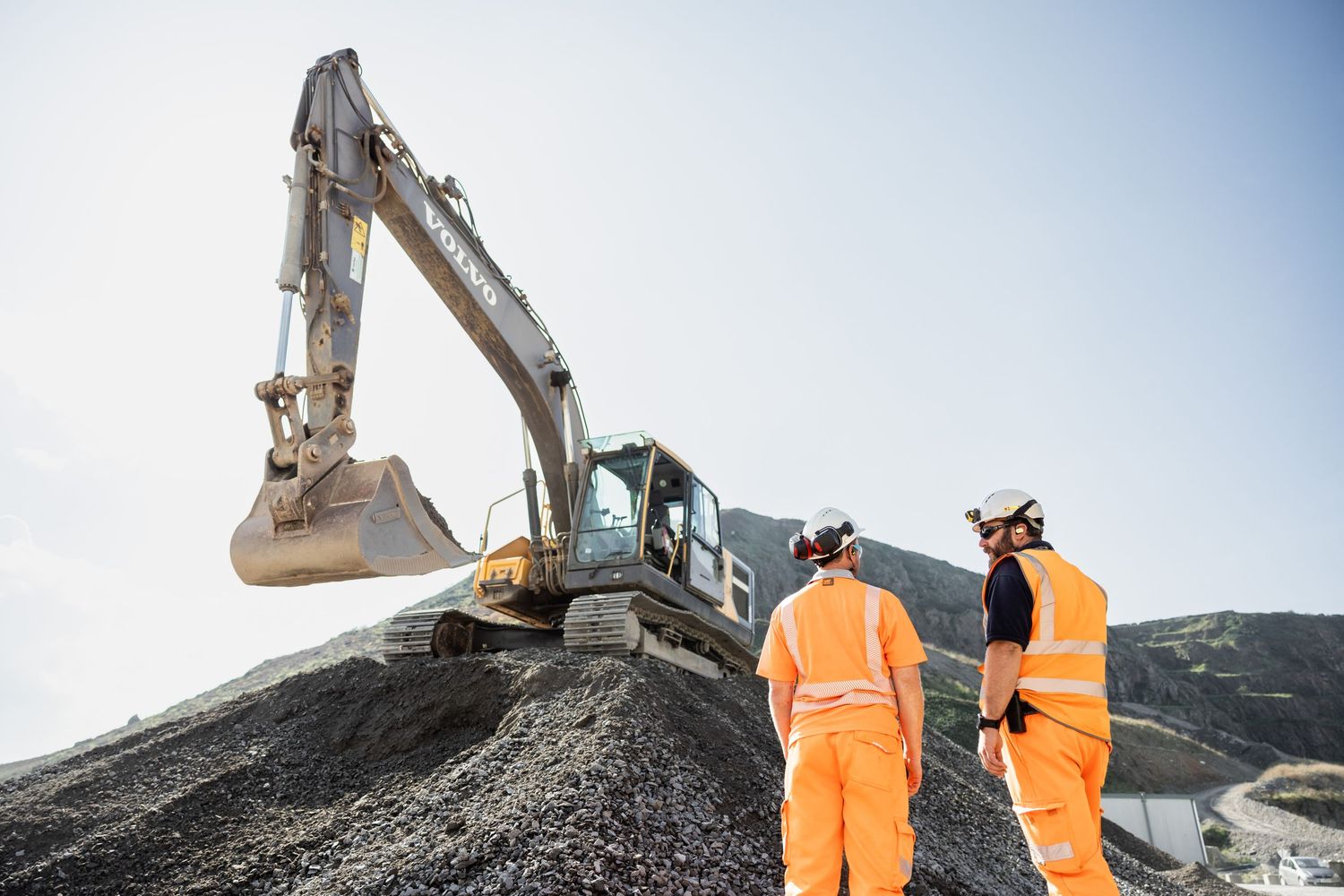 Construction workers in orange safety gear inspecting excavator at mining site.