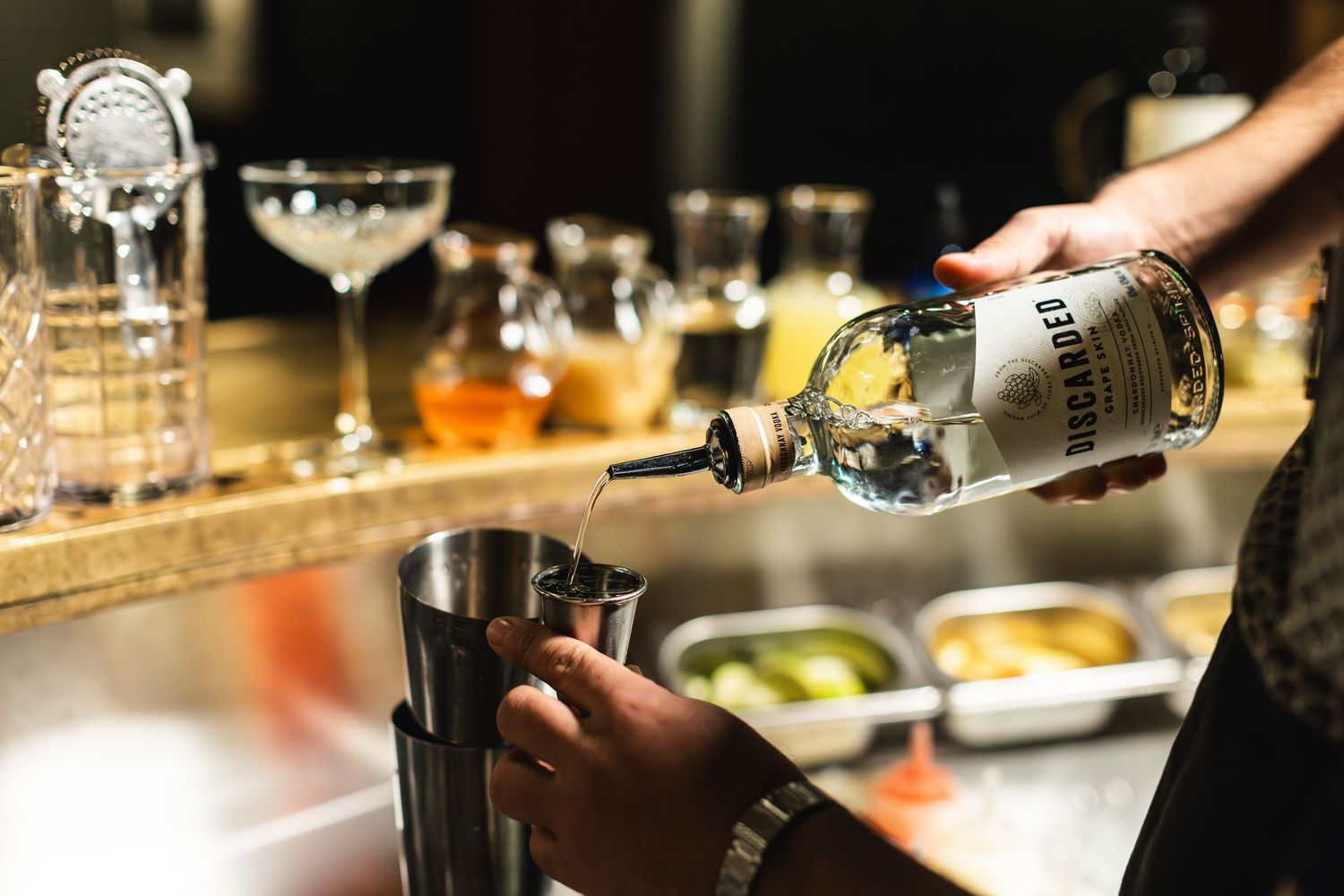 Hands pouring spirits into a cocktail shaker at a well-stocked bar.