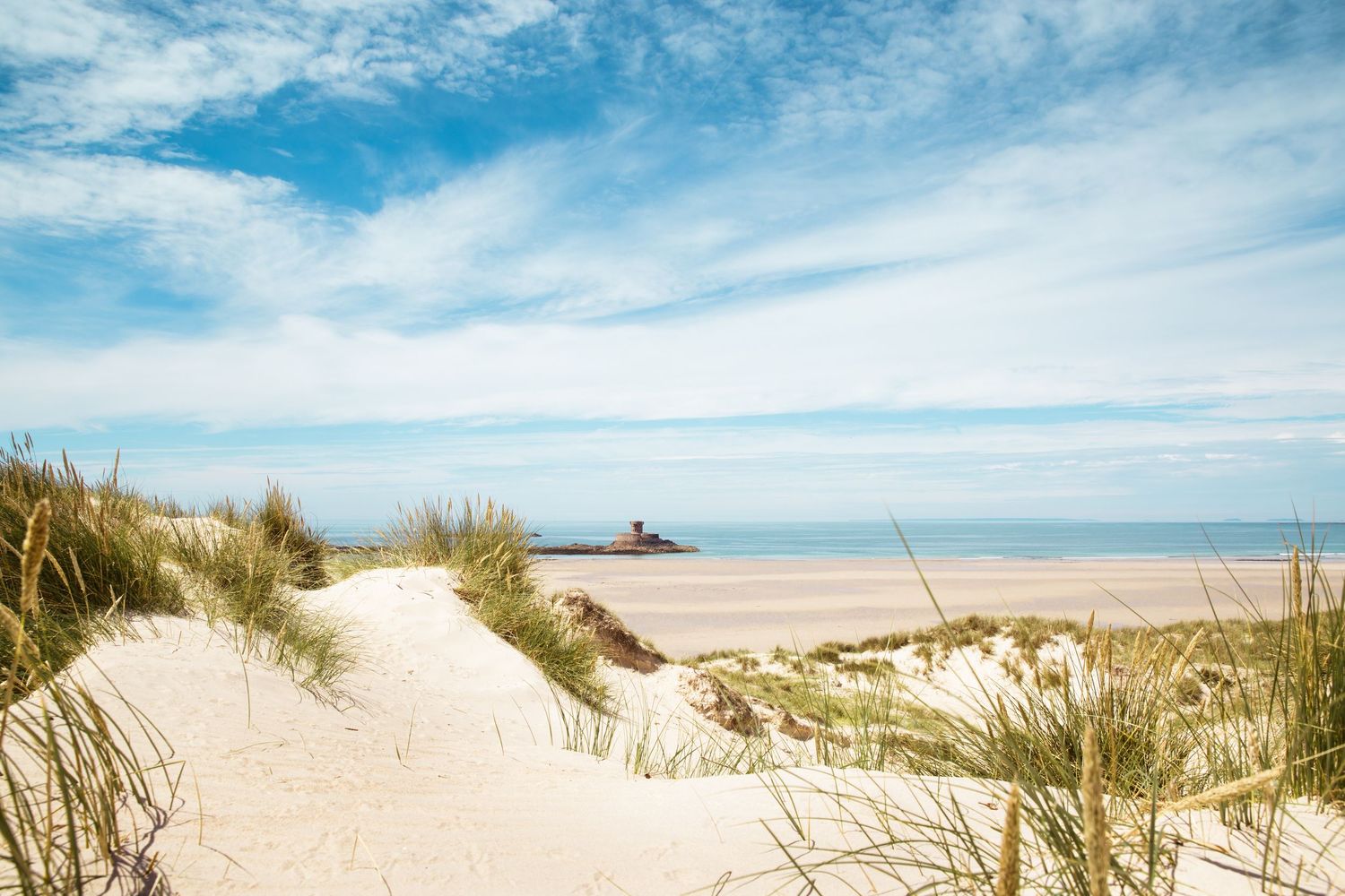 Sandy beach dunes with grass under bright blue sky and wispy clouds.