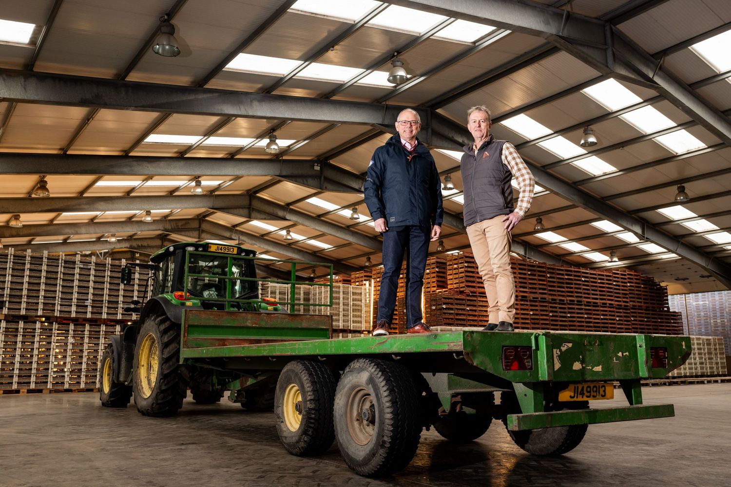 Two people standing on green John Deere farm equipment in industrial warehouse setting.