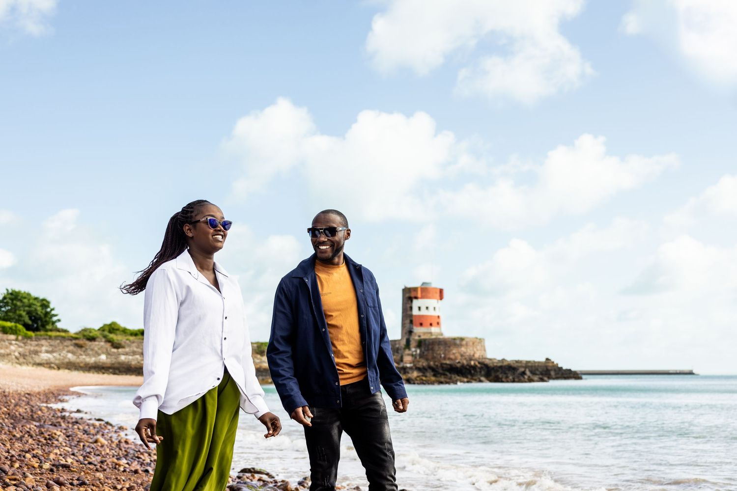 Couple walking along rocky shoreline with lighthouse and ocean views.