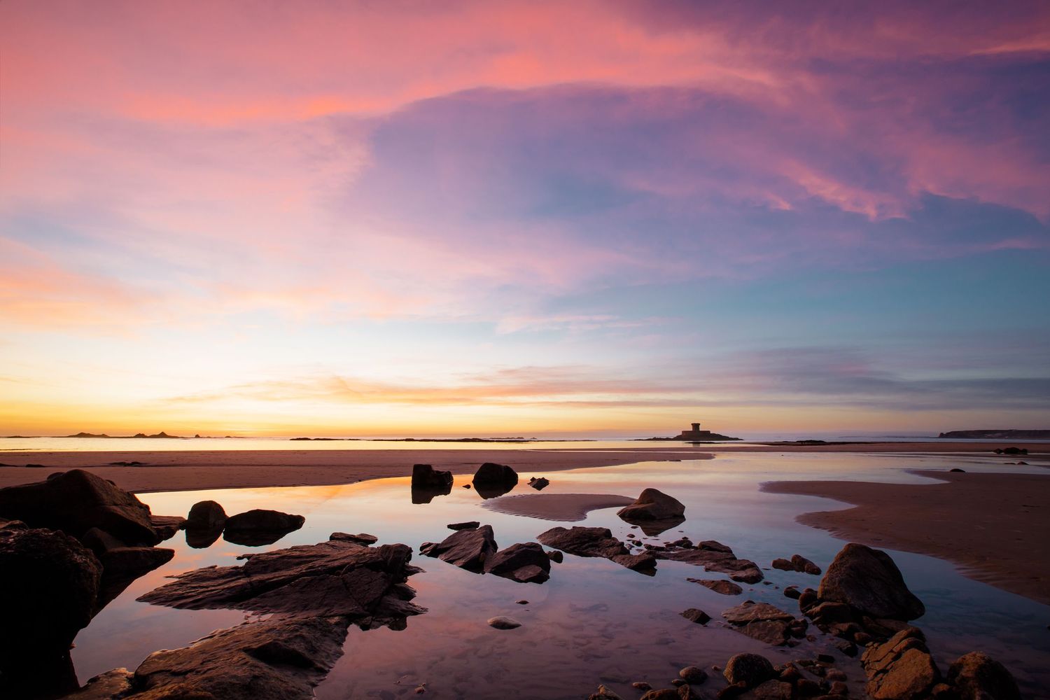 Dramatic sunset over rocky coastal shoreline with vibrant pink and purple clouds reflecting on calm ocean waters.