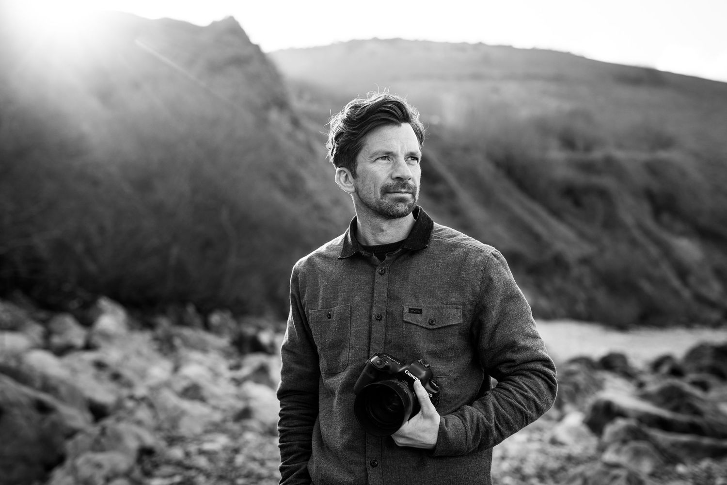 Portrait of a photographer holding camera equipment against a mountainous backdrop.