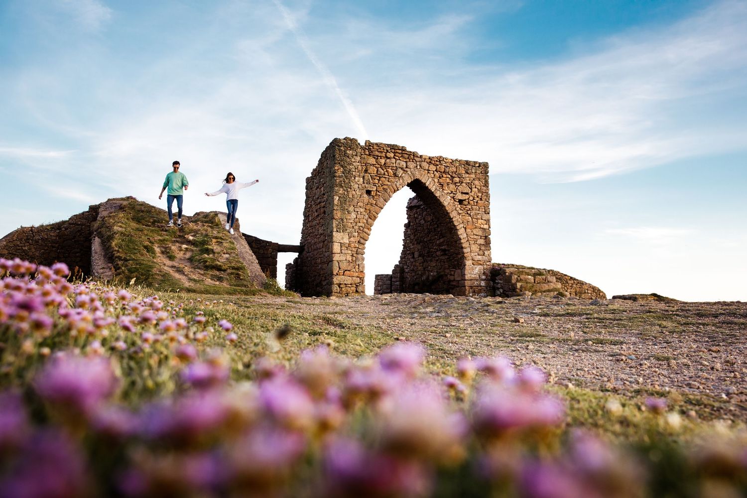 Ancient stone archway on a hilltop surrounded by pink wildflowers against a blue sky.
