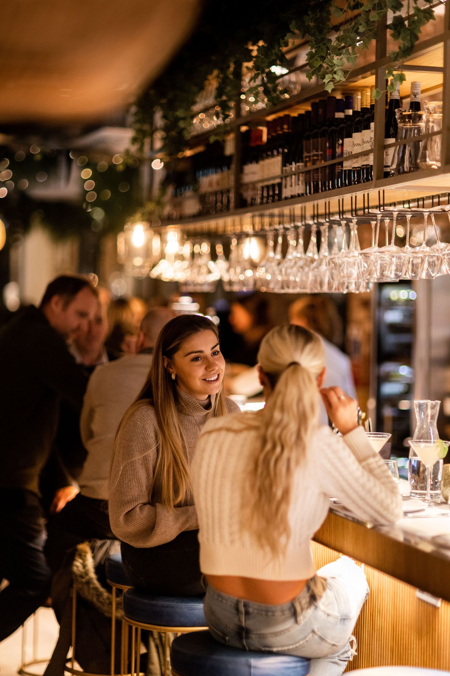 Patrons enjoying drinks at an elegant bar with ambient lighting and suspended glassware.