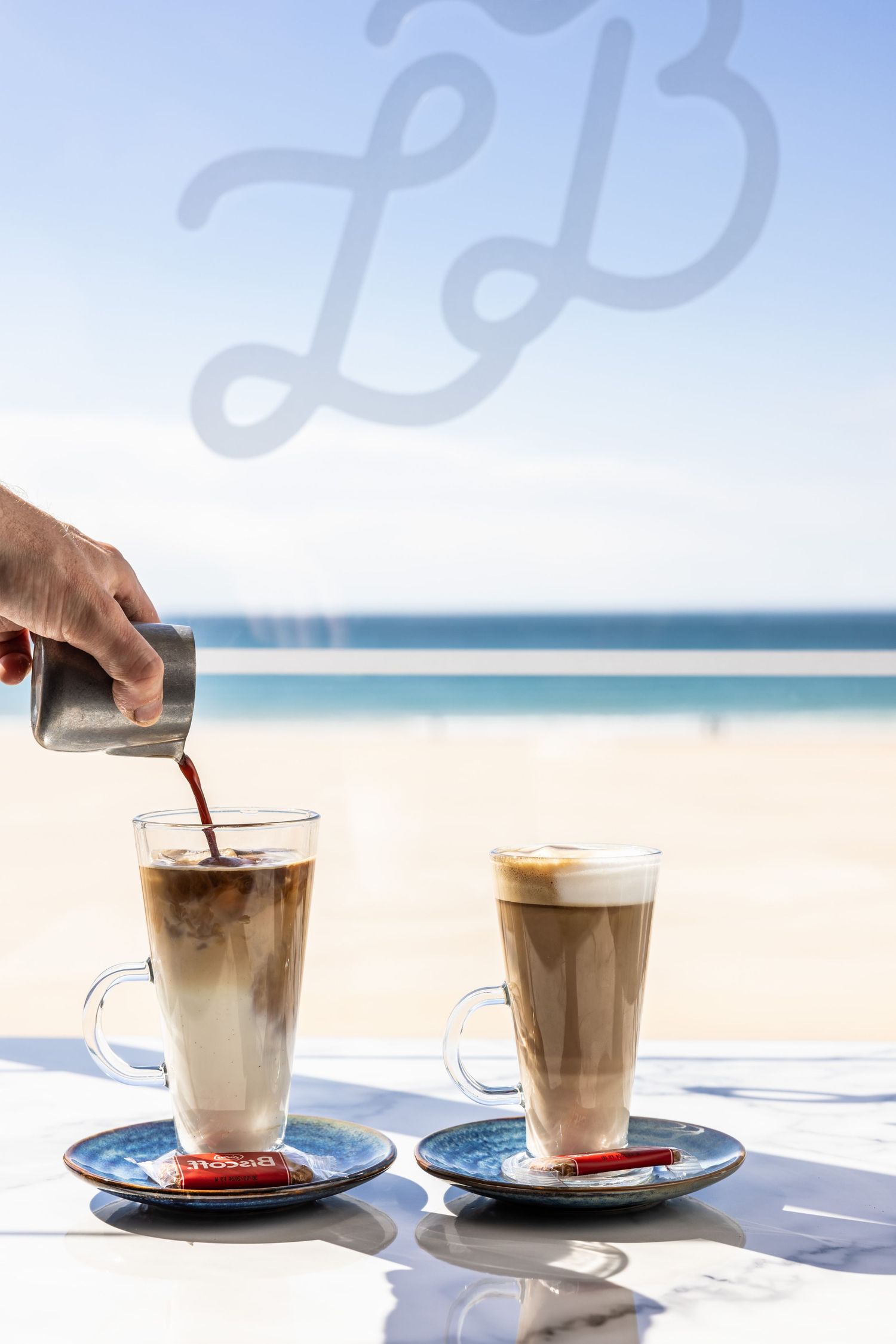 Two coffee drinks with layered foam served beachside with ocean view in the background.