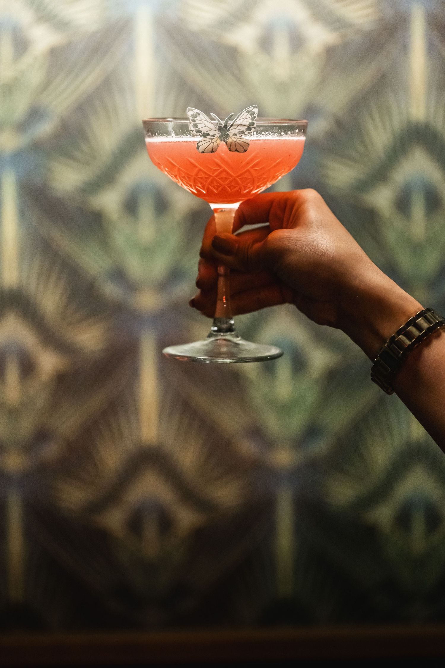 Hand holding a garnished red cocktail against a decorative art deco wallpaper background.