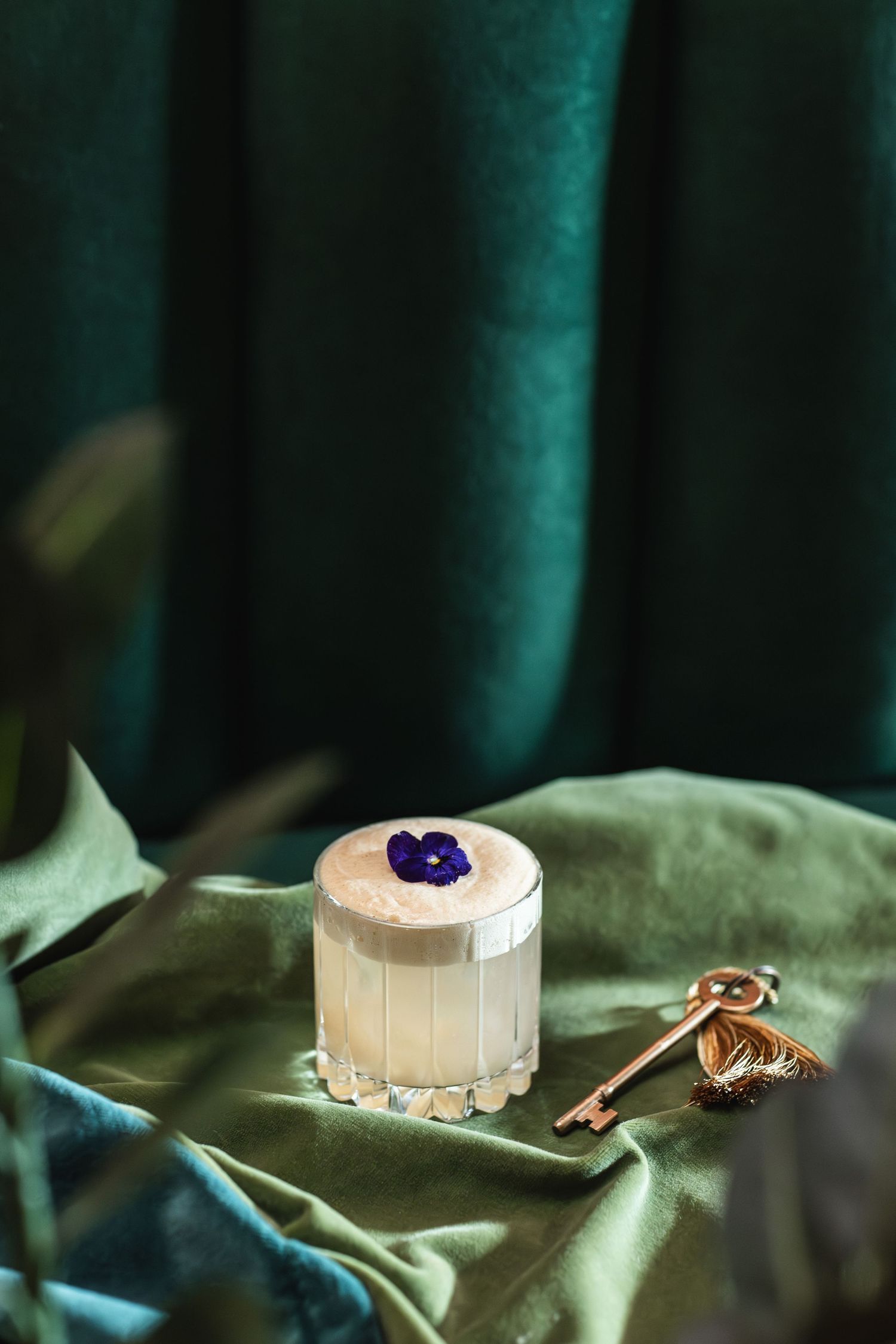 A creamy dessert cocktail with a blue garnish and vintage key on emerald velvet.