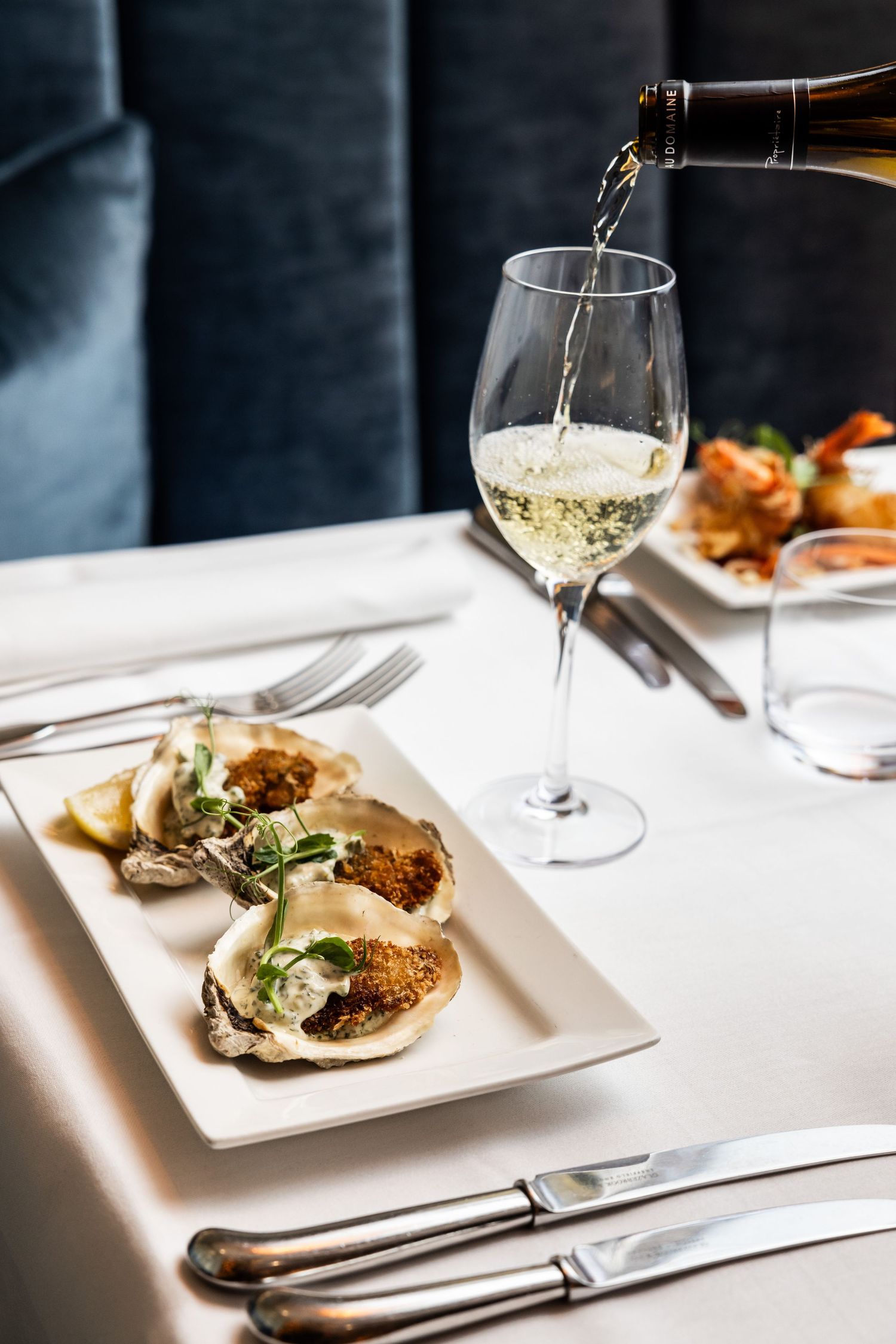 White wine being poured over fresh oysters on a pristine restaurant table setting.