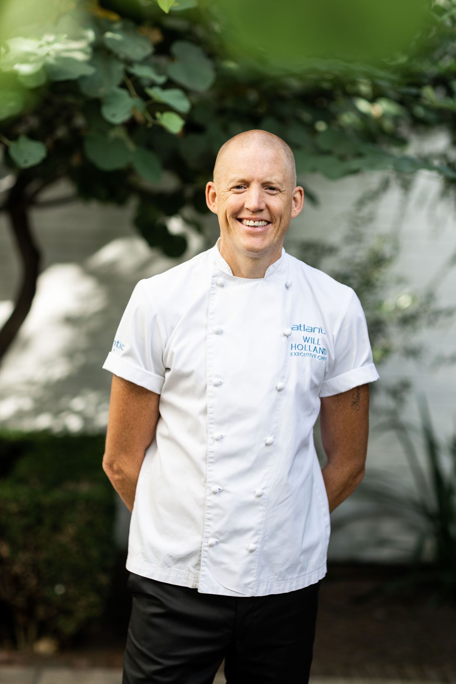 Professional chef in white uniform smiling outdoors in natural garden setting.