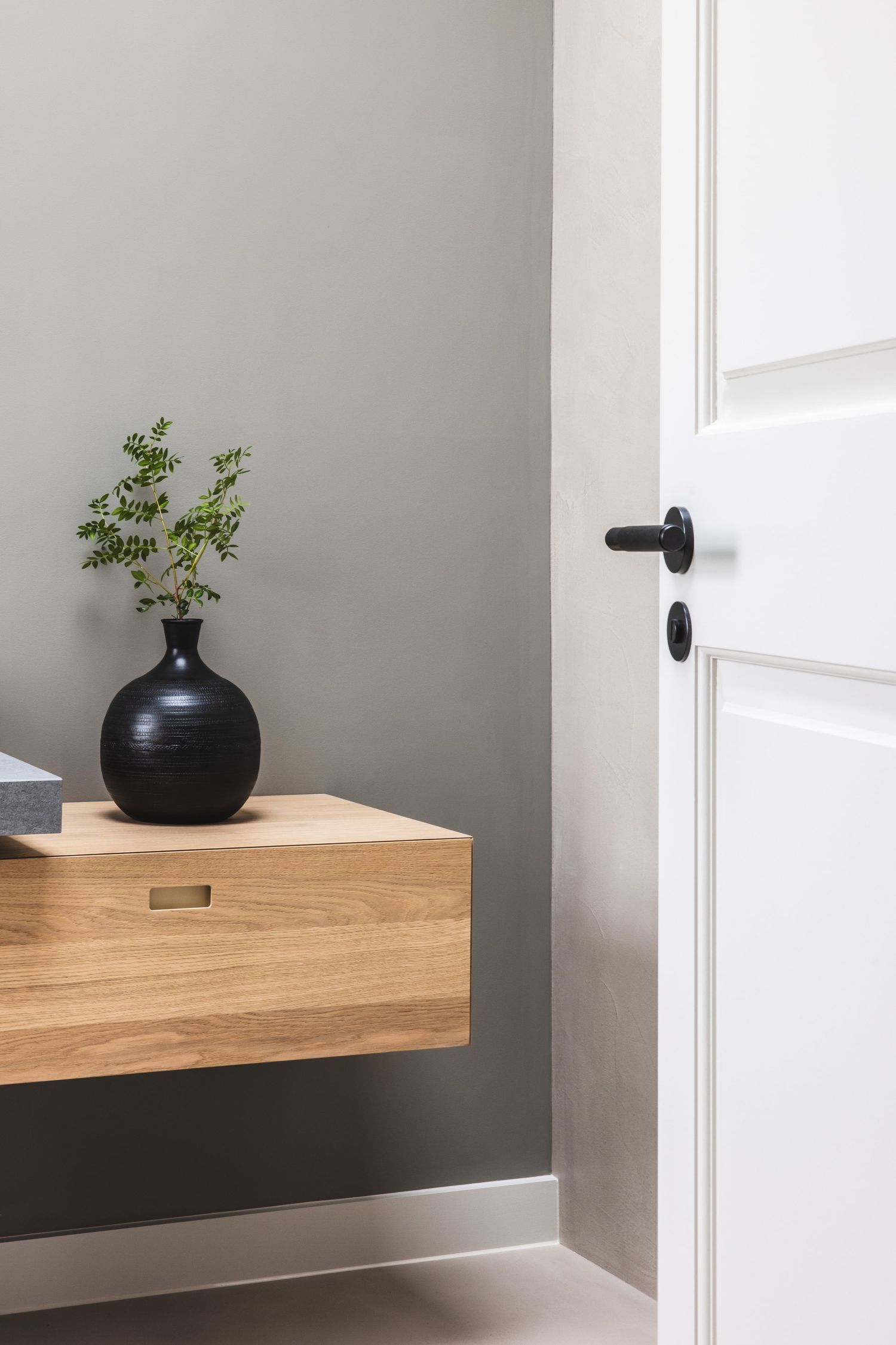Floating wooden shelf with black vase and plant against gray wall in minimalist bathroom.