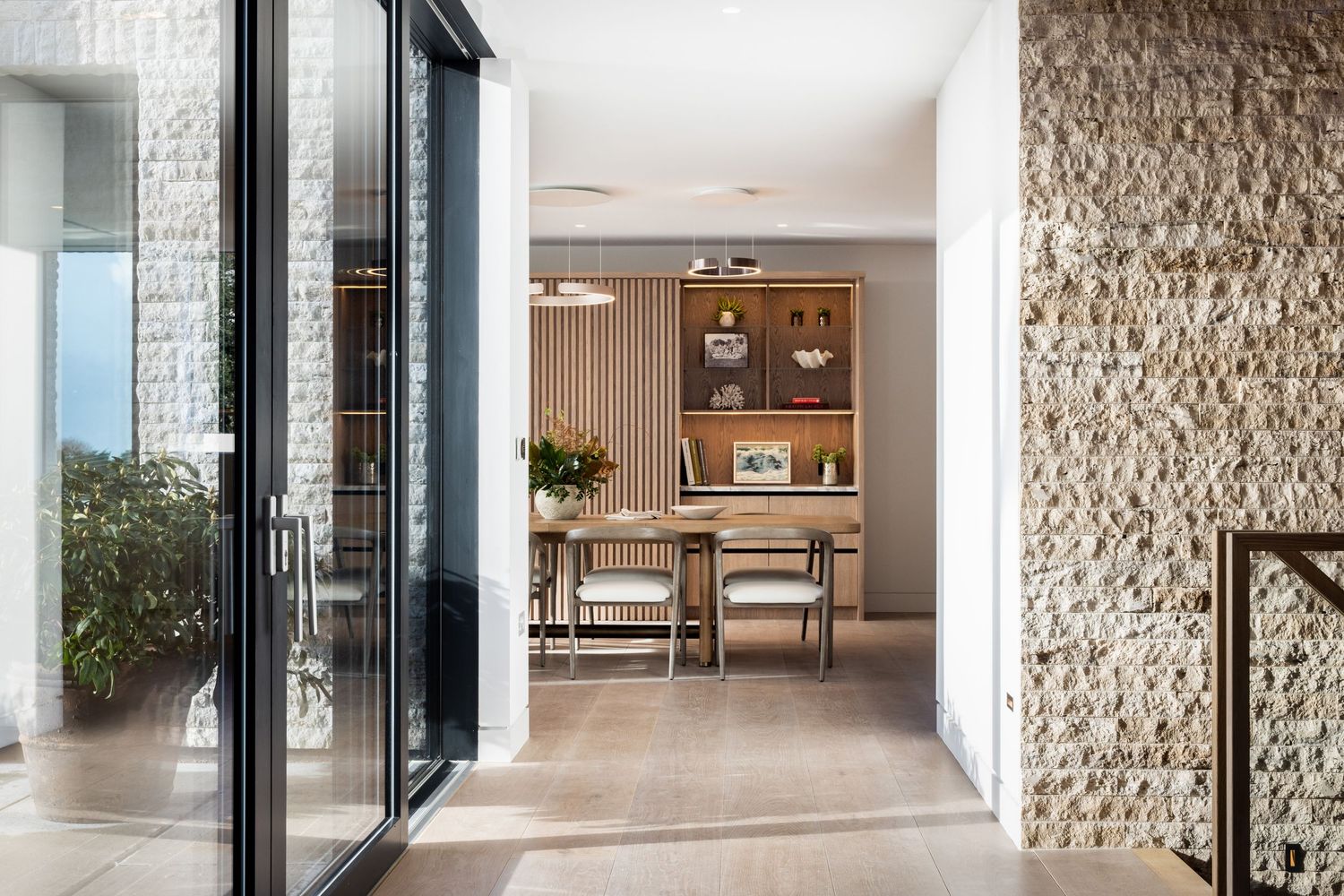 Contemporary hallway with glass sliding doors, stone wall texture, and wooden dining area in background.