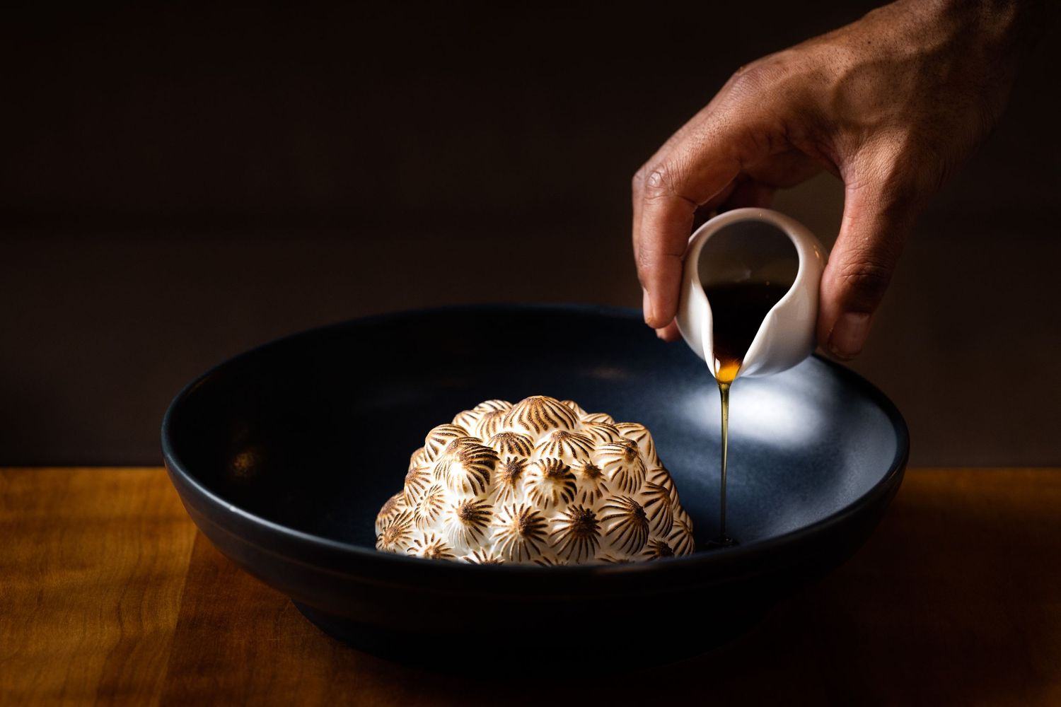 Hand pouring sauce over a textured baked dessert in a black bowl.