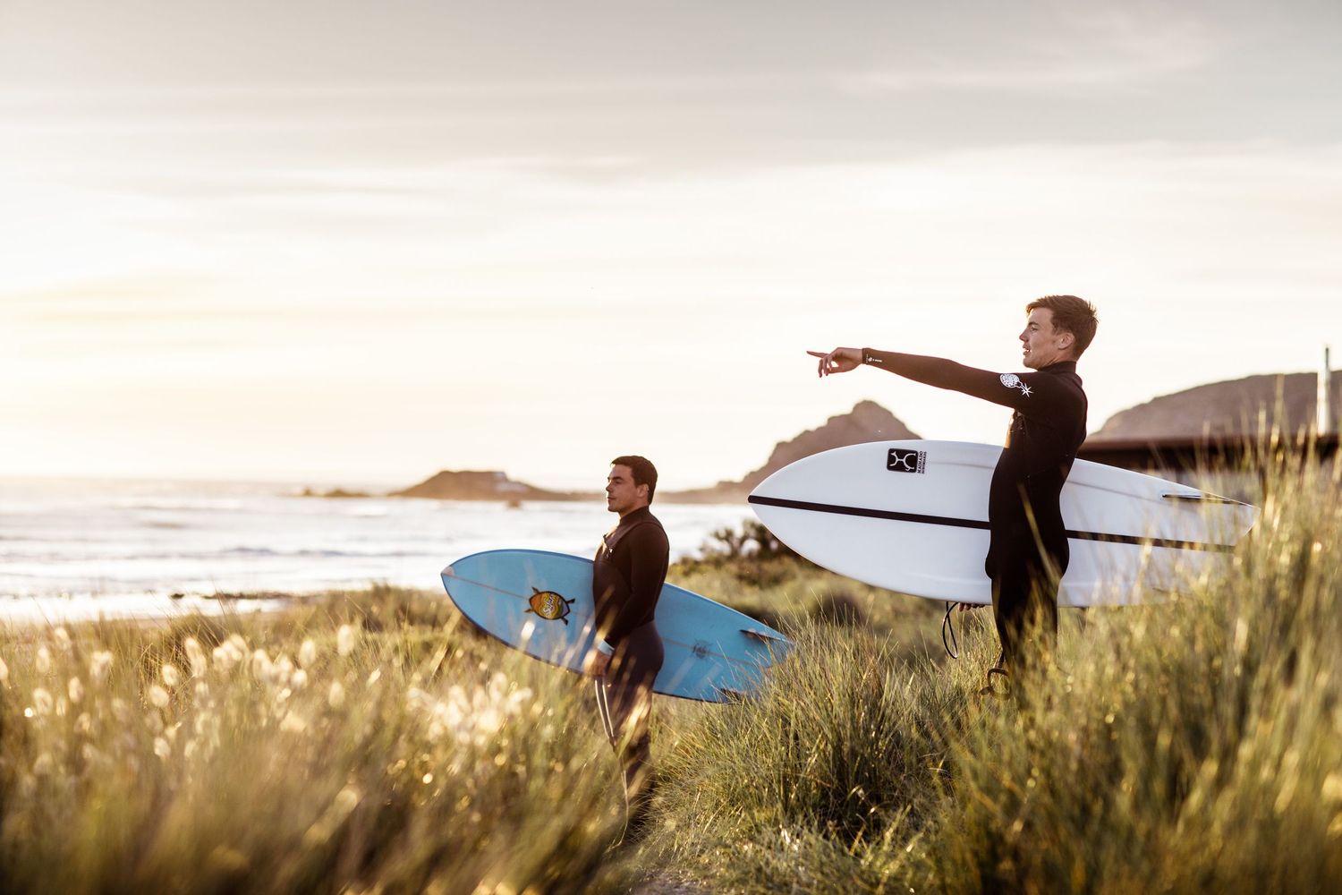Two surfers carrying boards through tall grass at sunset near the beach.